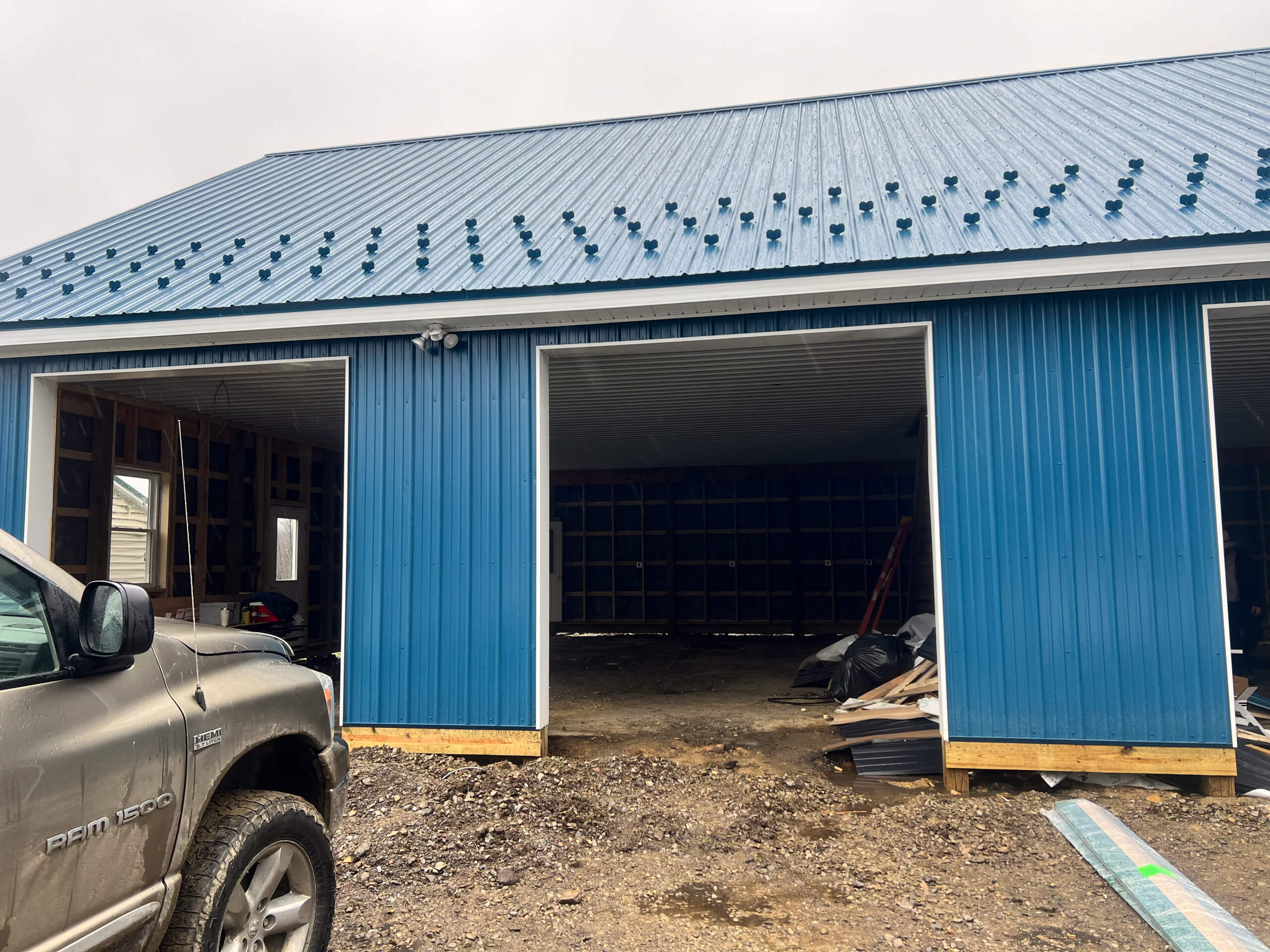 Crawford County Roofing Spartasburg PA A blue metal building under construction with two large open garage doors, construction materials on the ground, and a parked truck in front.