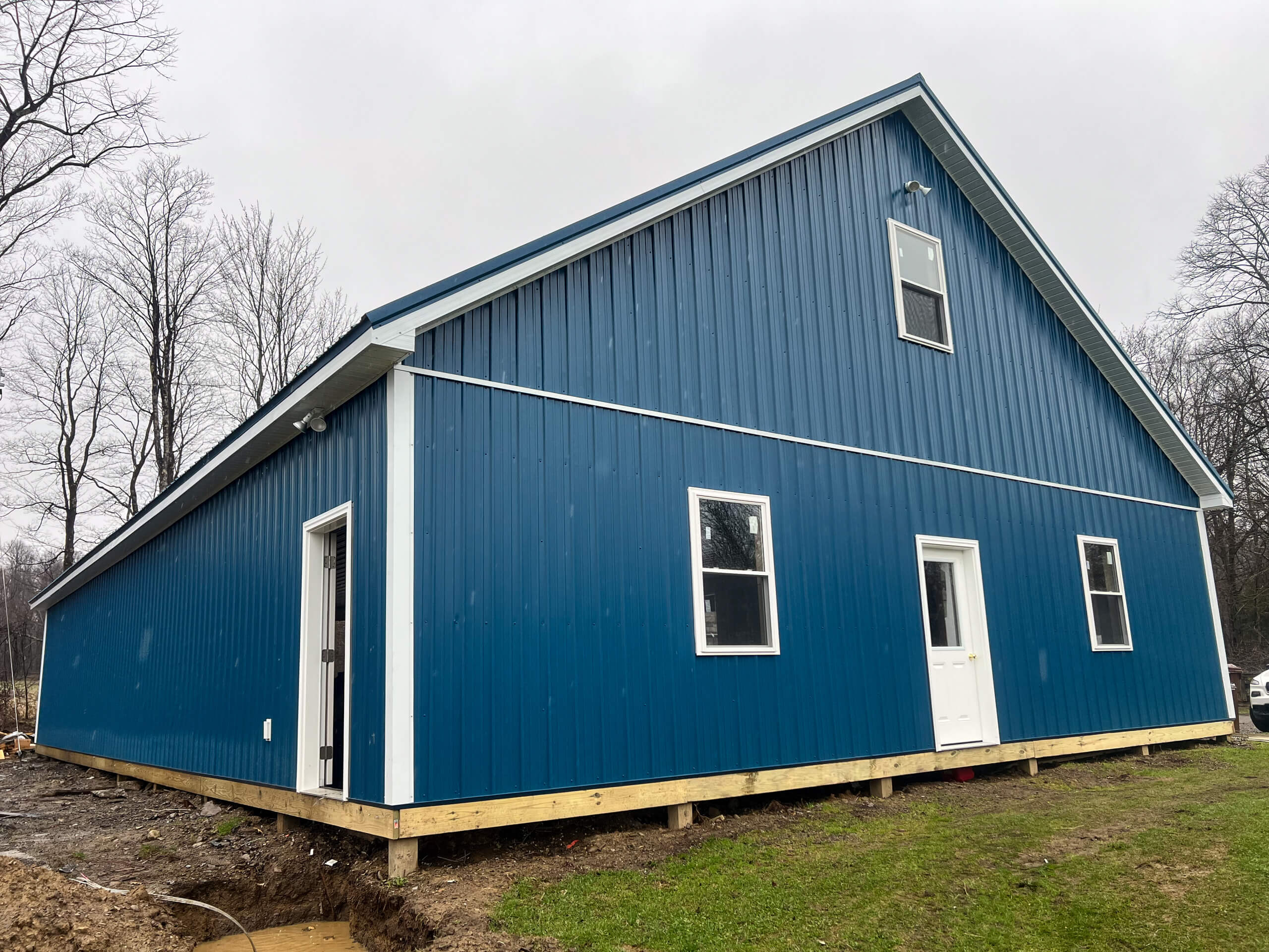 Crawford County Roofing Spartasburg PA A blue metal-sided building with white trim and doors sits on a raised foundation, surrounded by bare trees and grass under an overcast sky.