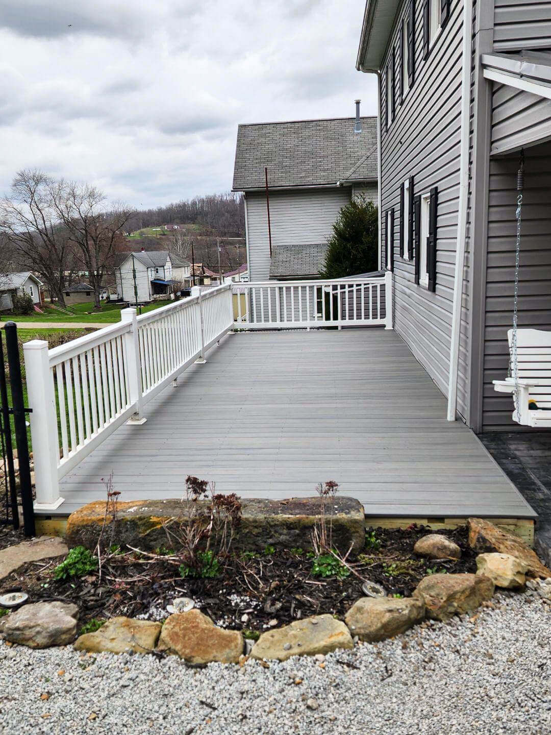 A gray wooden deck with white railings attached to a gray house, overlooking a small garden bed bordered by rocks and a gravel area.