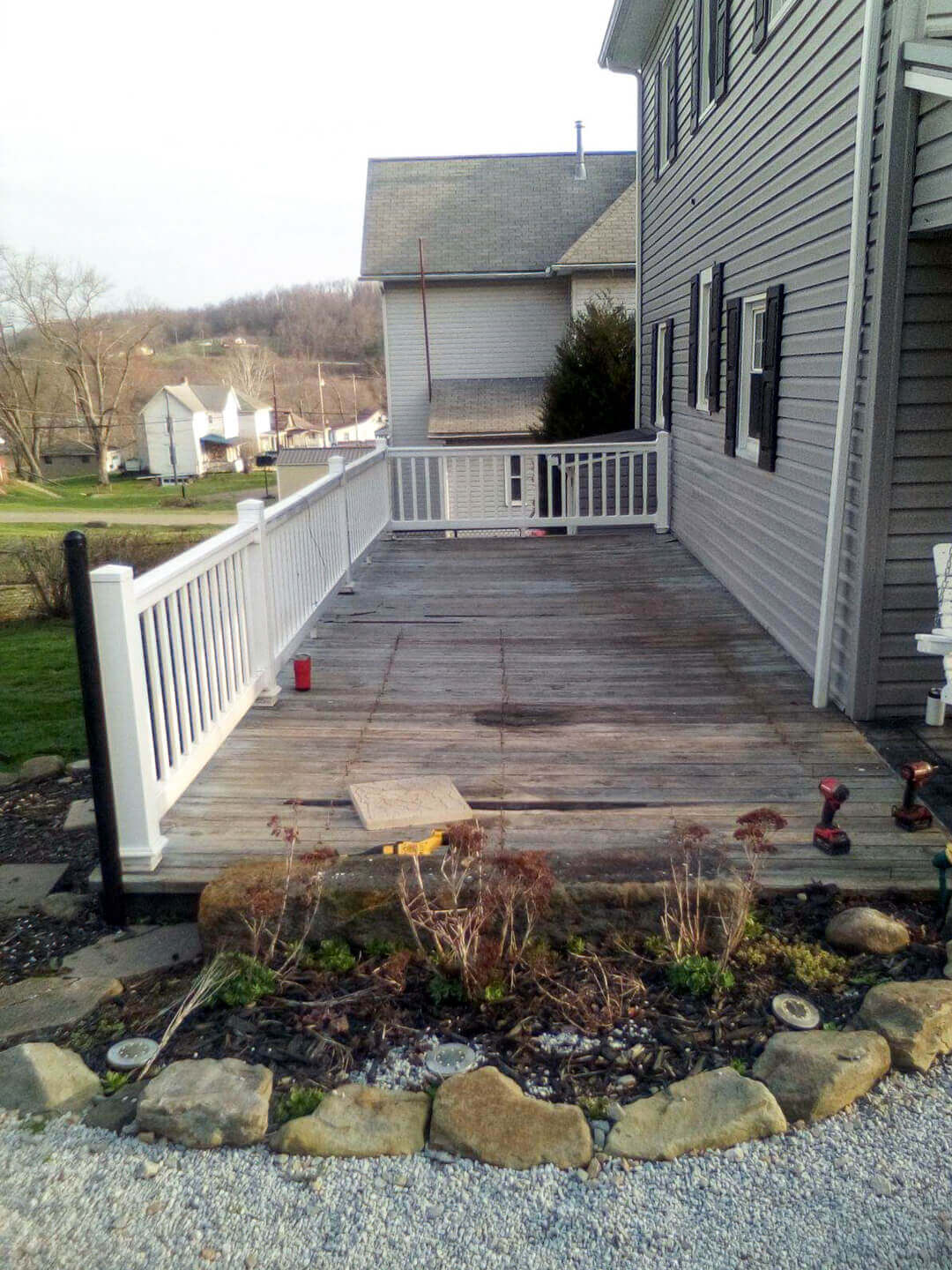 A weathered wooden deck with white railings attached to a house, overlooking a small garden with rocks and dried plants; tools and a red cup sit on the deck.