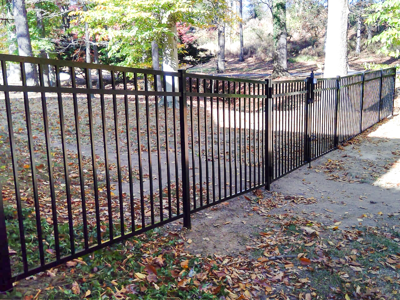 A black metal fence with a gate stands open across a leaf-covered path in a wooded area during autumn.