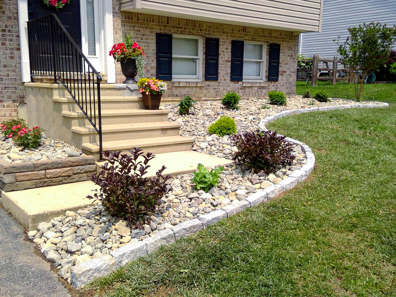 Front yard with stone landscaping, small shrubs, and flowers along a curved border next to a concrete walkway and steps leading to a brick house with black shutters.