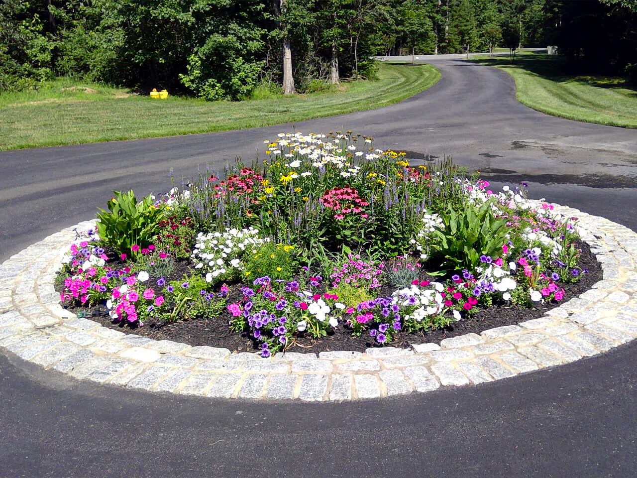 A circular flower bed with various colorful flowers in the center of a roundabout, bordered by a stone edge and surrounded by a paved road with trees and grass in the background.