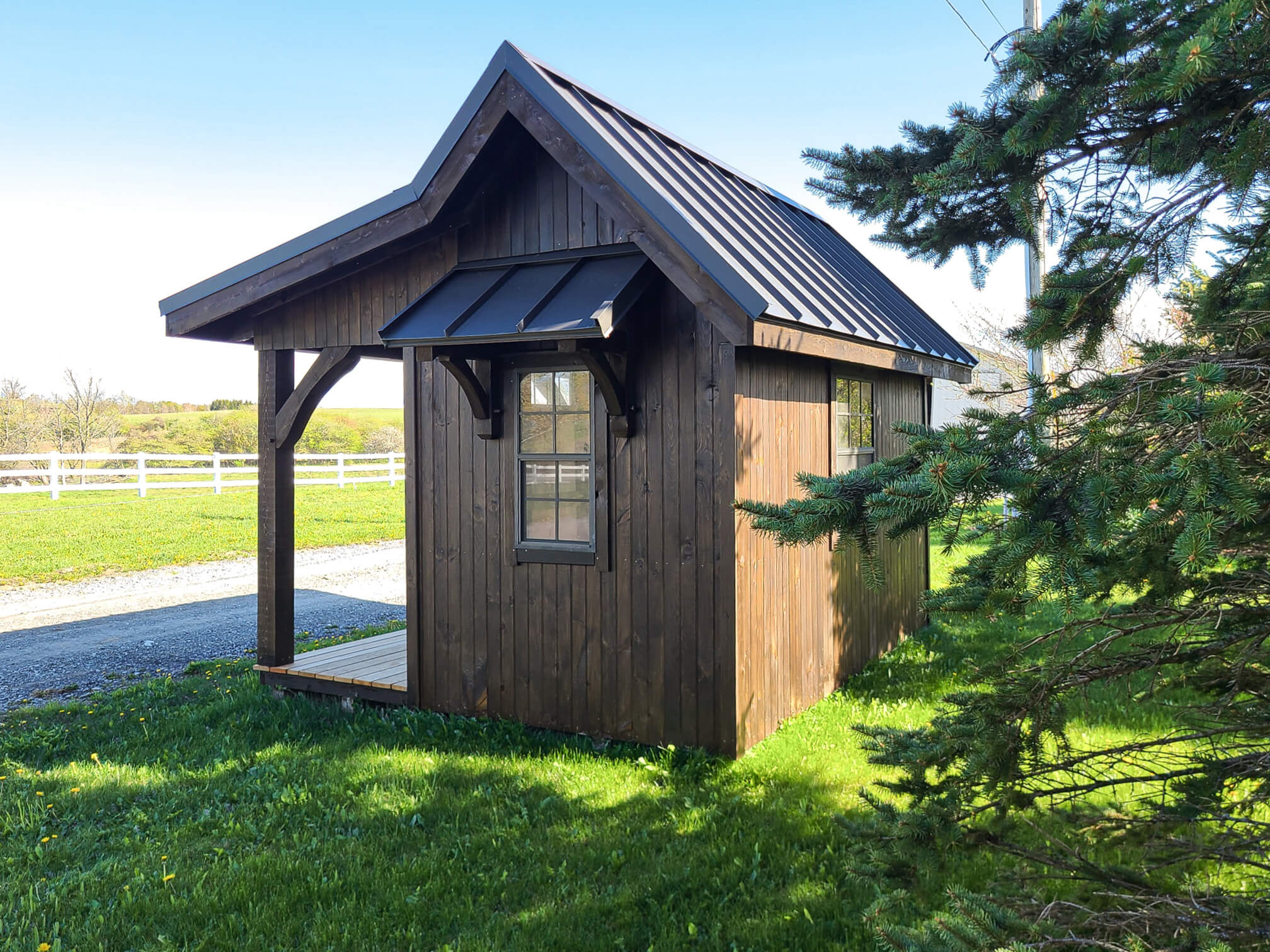 Elite Timberframes Canastota, NY A small, dark brown wooden shed with a metal roof sits on grass near a gravel path, with a pine tree and white fencing in the background.