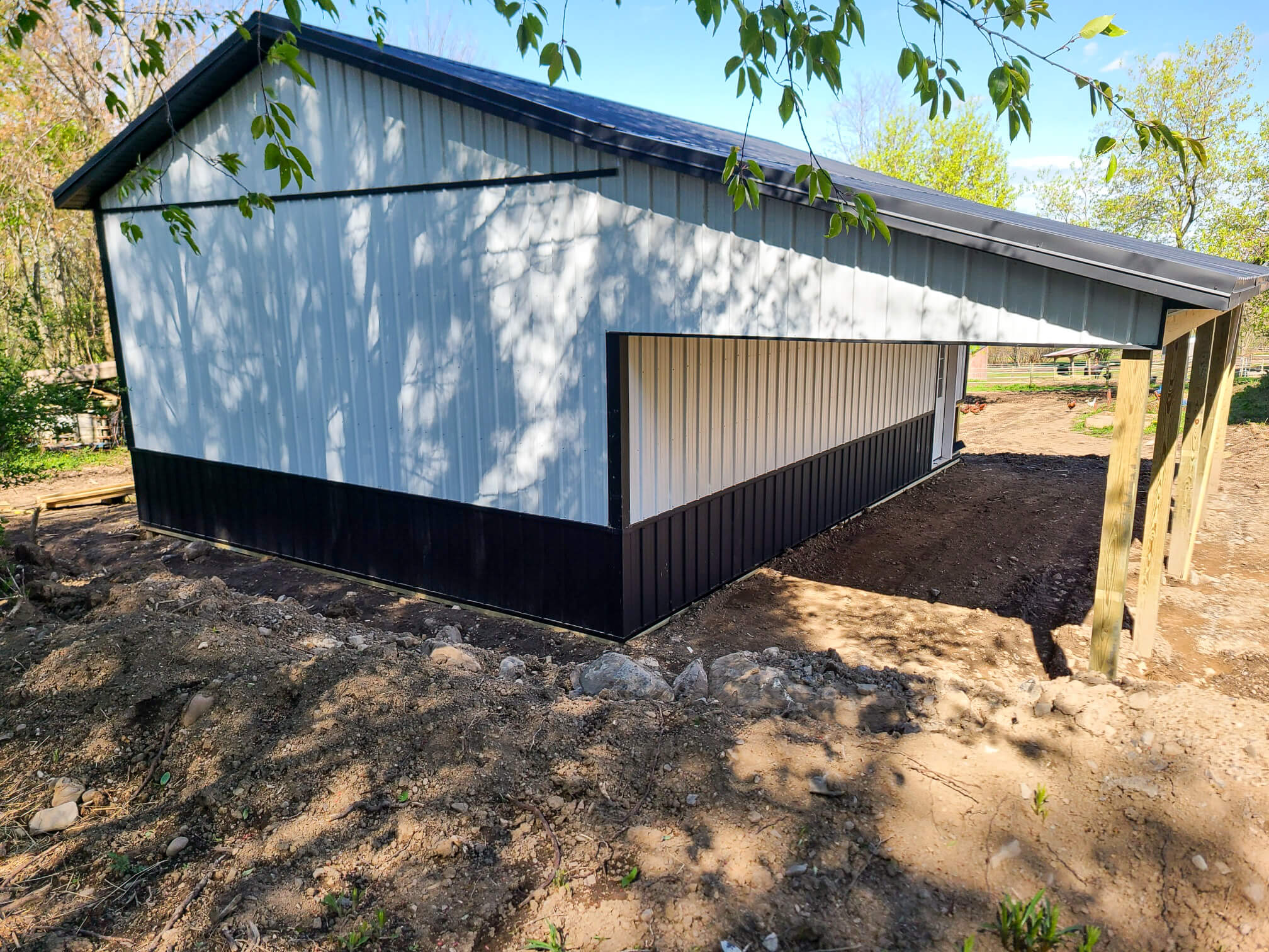 Elite Timberframes Canastota, NY A newly constructed metal-sided shed with a sloped roof stands on a dirt lot surrounded by trees and partially shaded by branches.