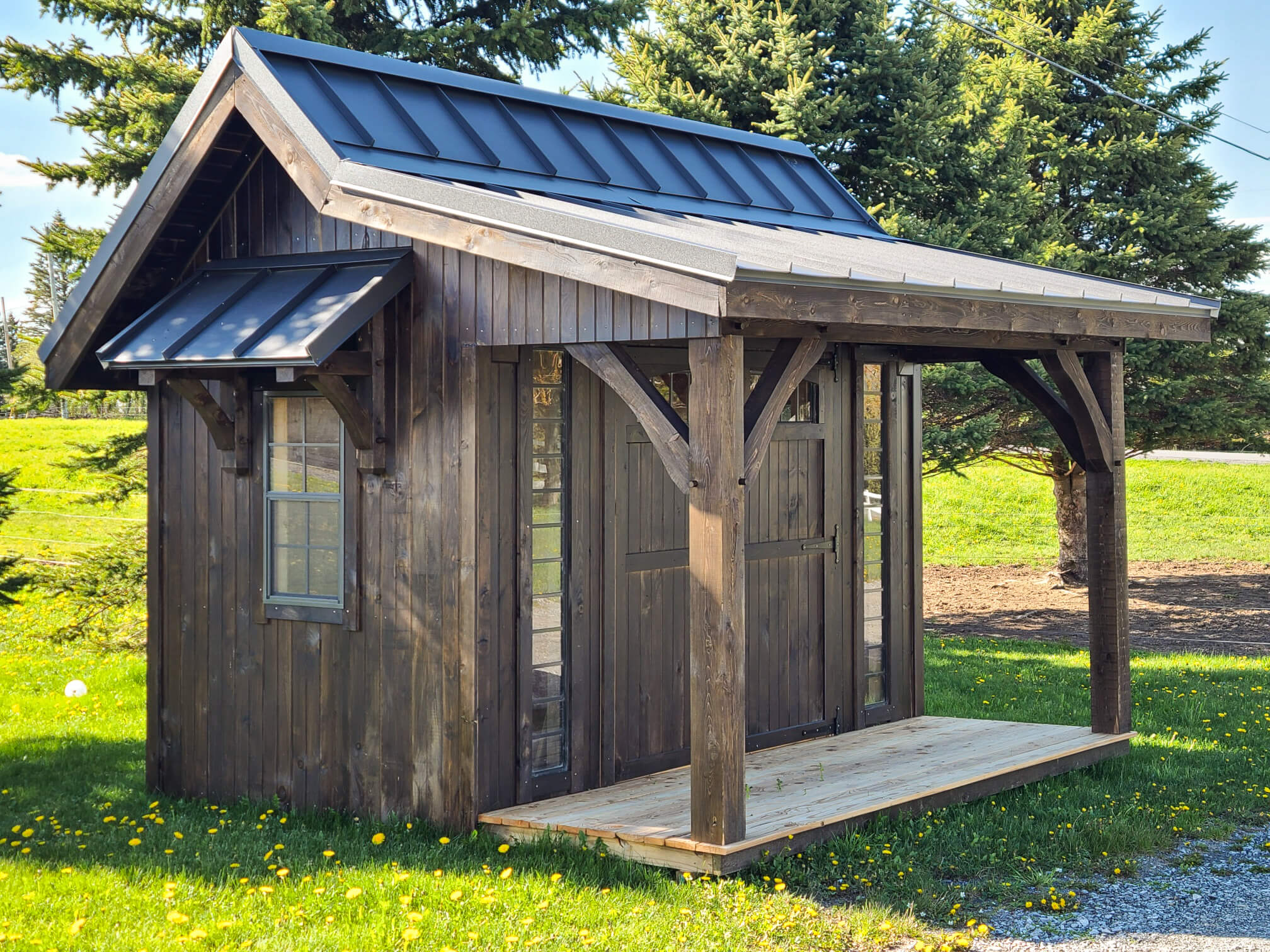 Elite Timberframes Canastota, NY A small wooden shed with a metal roof and a covered porch stands on a grassy lawn with trees in the background.