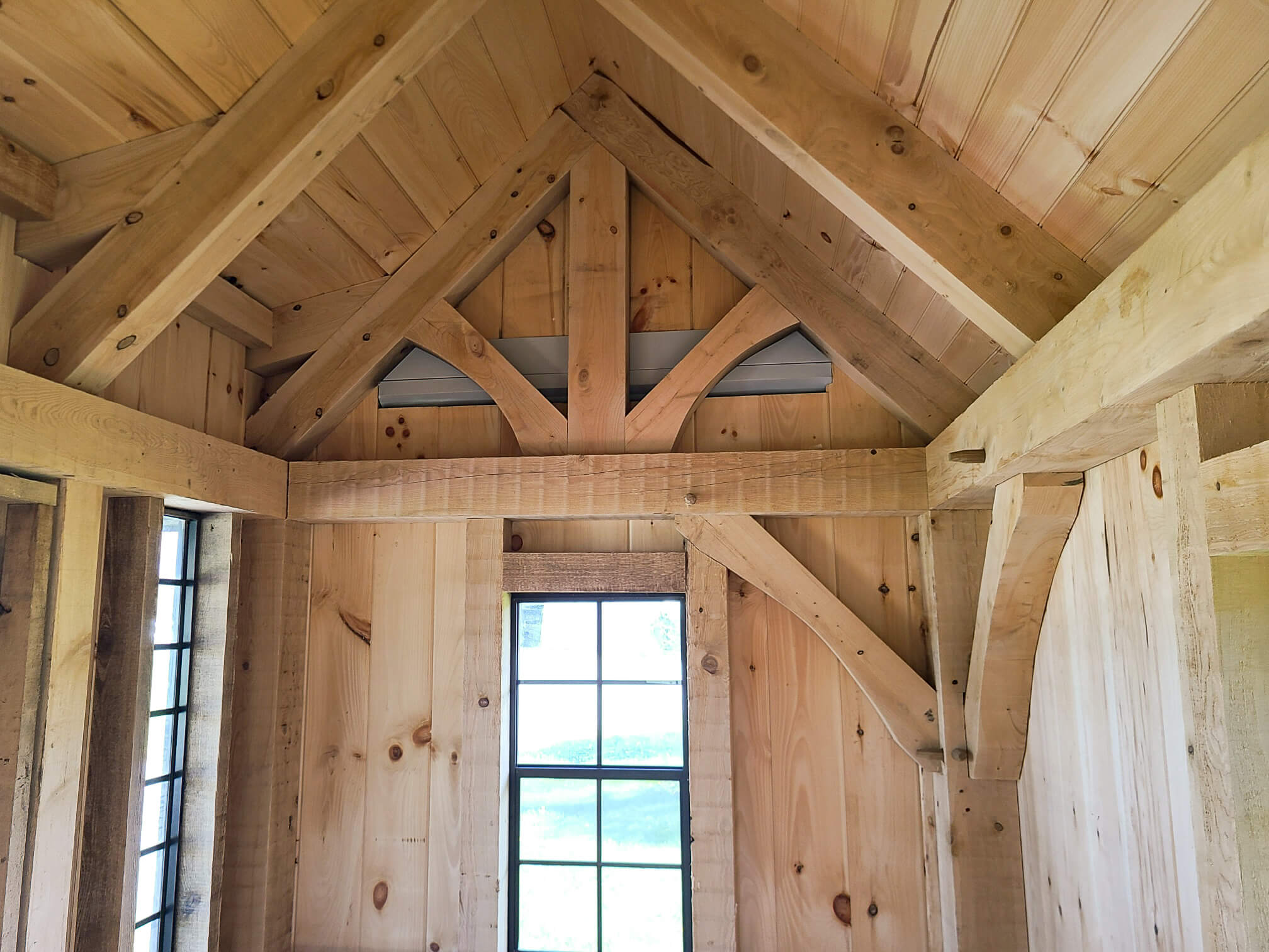 Elite Timberframes Canastota, NY Interior of a wooden structure showing exposed beams, vaulted ceiling, and two vertical windows letting in natural light.