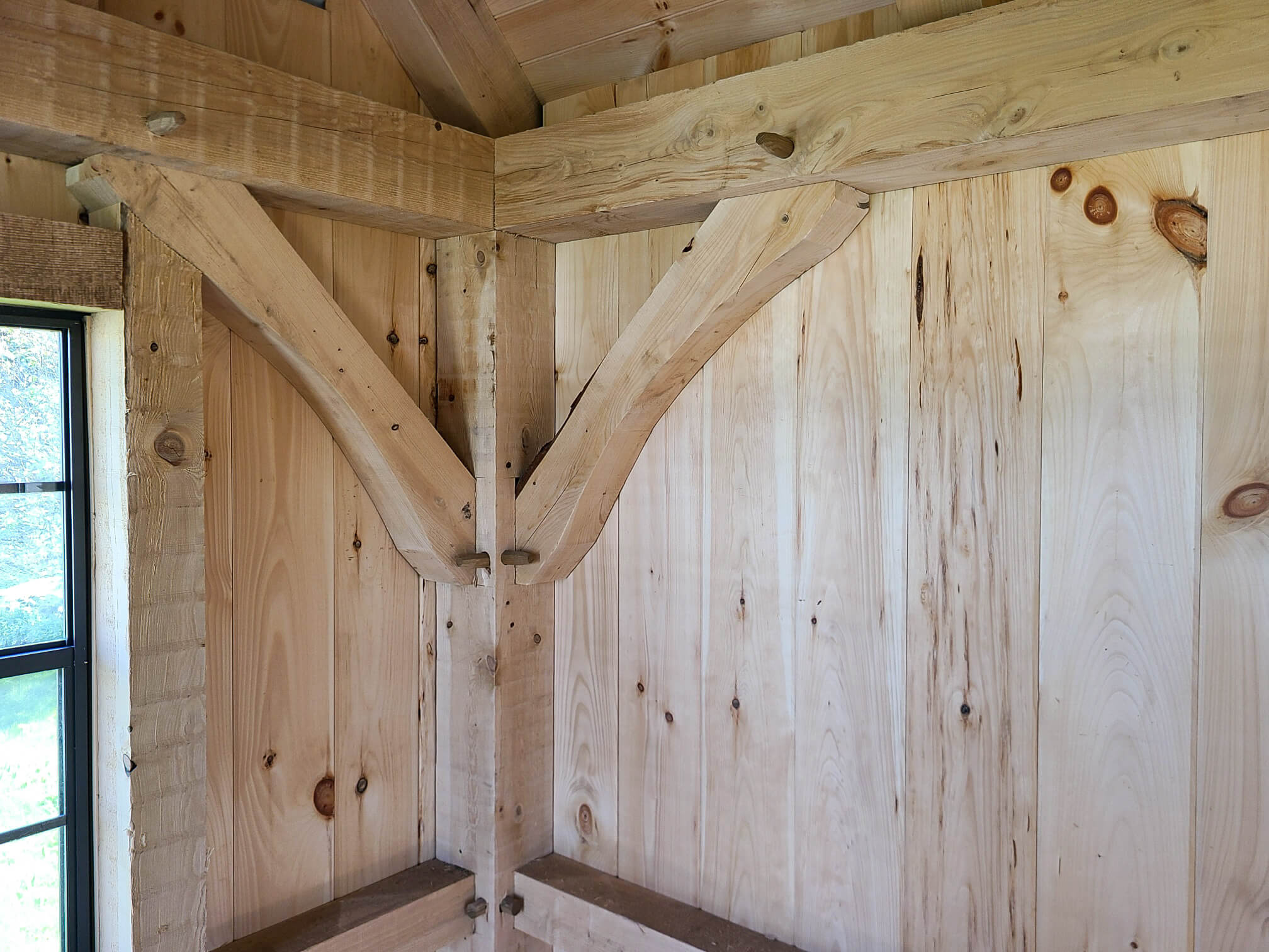 Elite Timberframes Canastota, NY Wooden interior corner showing timber frame joinery with exposed beams, braces, and pegs next to a window with sunlight coming in.