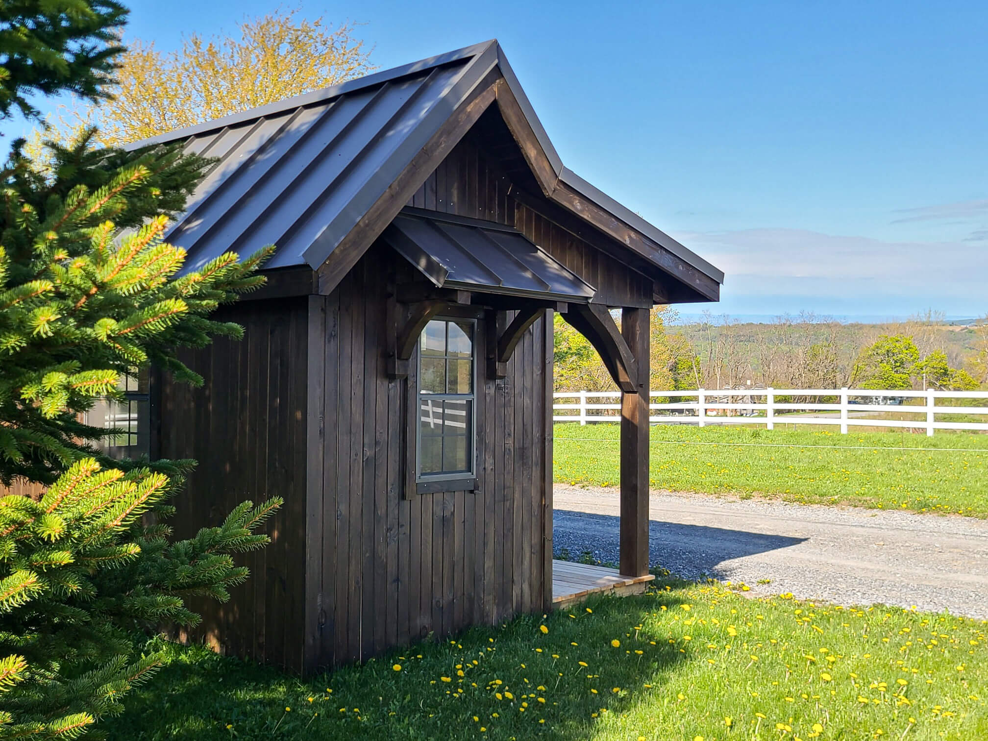 Elite Timberframes Canastota, NY A small wooden shed with a metal roof stands beside a gravel path, surrounded by grass, dandelions, a pine tree, and a white fence under a clear blue sky.
