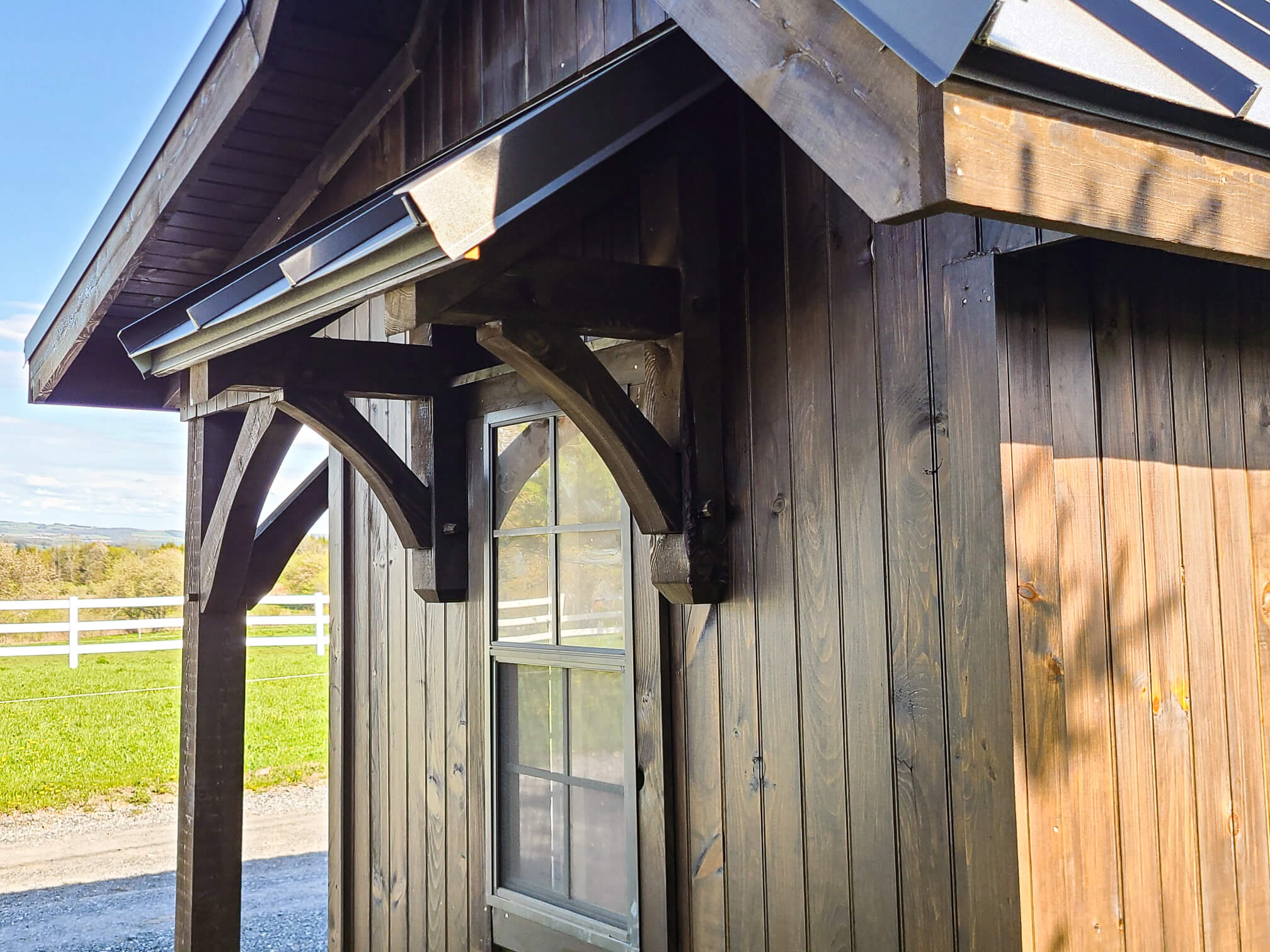 Elite Timberframes Canastota, NY A close-up of a wooden shed with a window and overhanging roof, set near a green field and white fence under a clear sky.