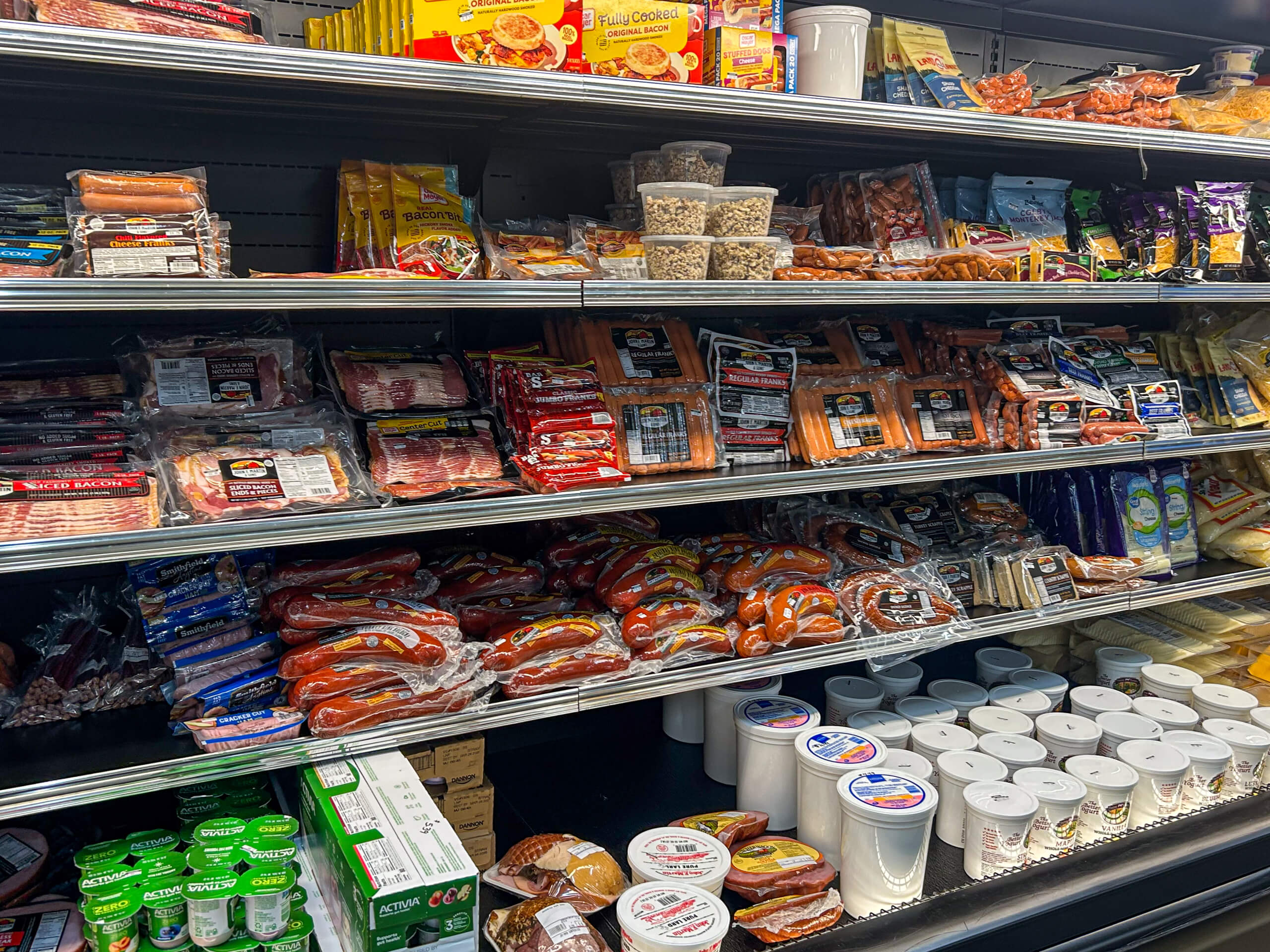 Jamar Groceries Kutztown PA Refrigerated grocery shelves stocked with packaged meats, sausages, hot dogs, dairy products, various cheeses, and containers of seeds or nuts.