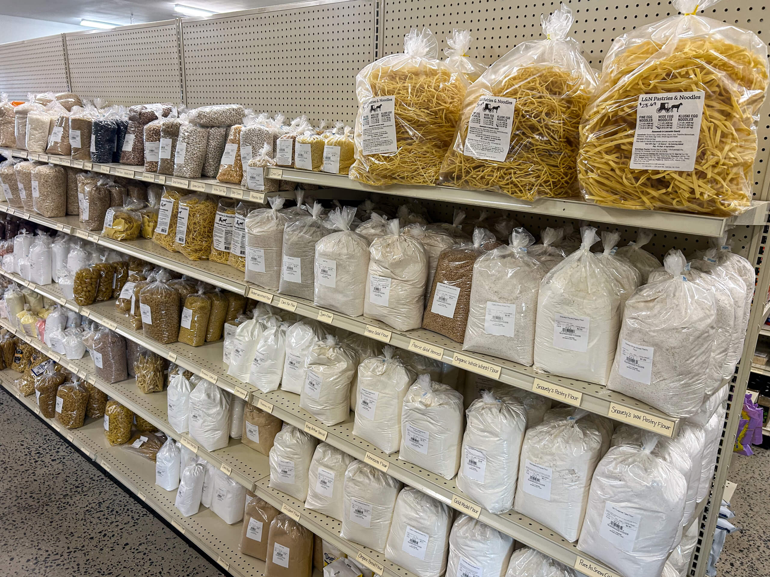 Jamar Groceries Kutztown PA Shelves in a store display various packaged dry goods, including different types of noodles, beans, and flours in clear plastic bags with labels.