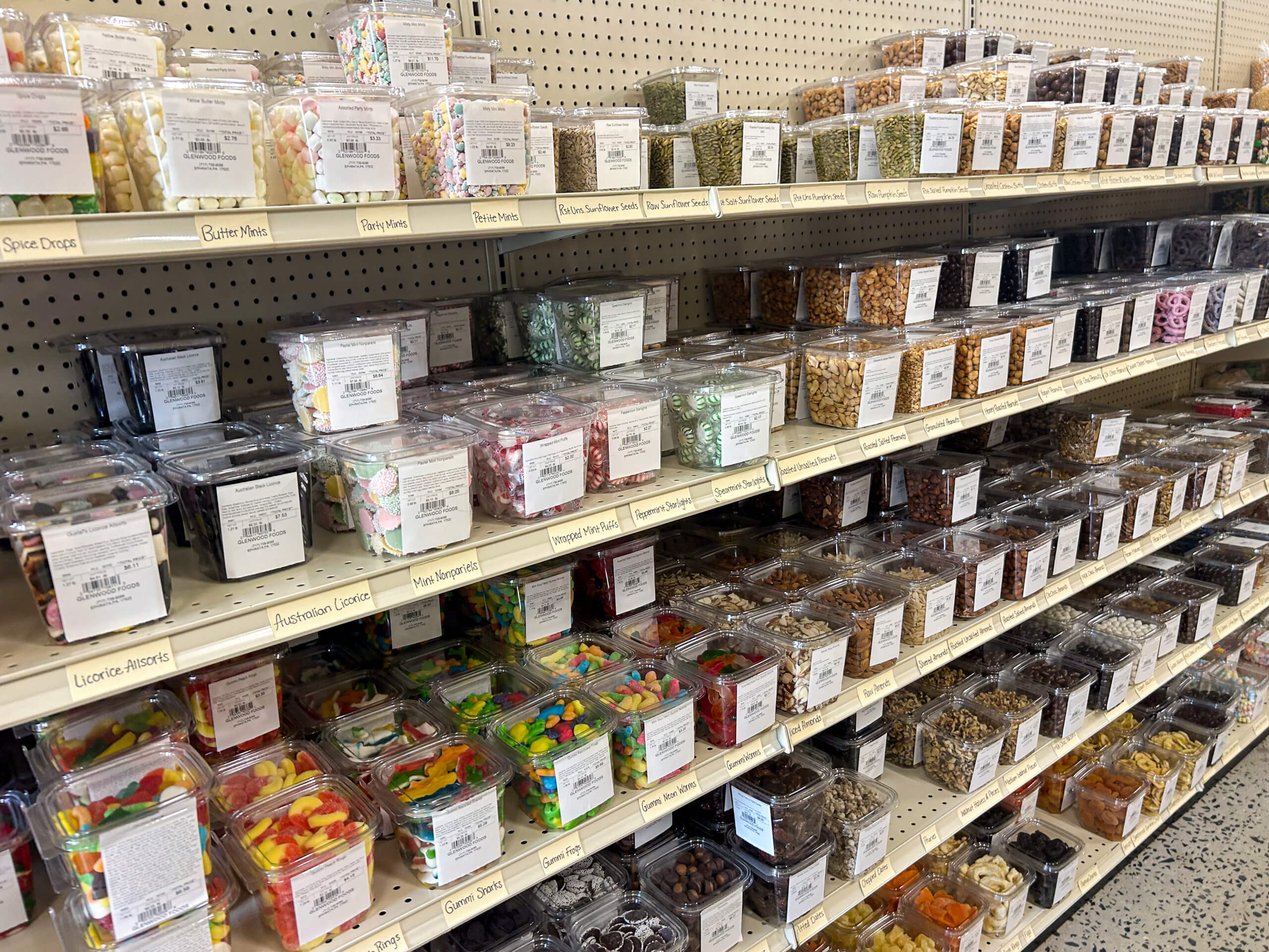 Jamar Groceries Kutztown PA Shelves display a variety of bulk candies, nuts, and snacks in clear plastic containers, each labeled with product and price information at a grocery store.