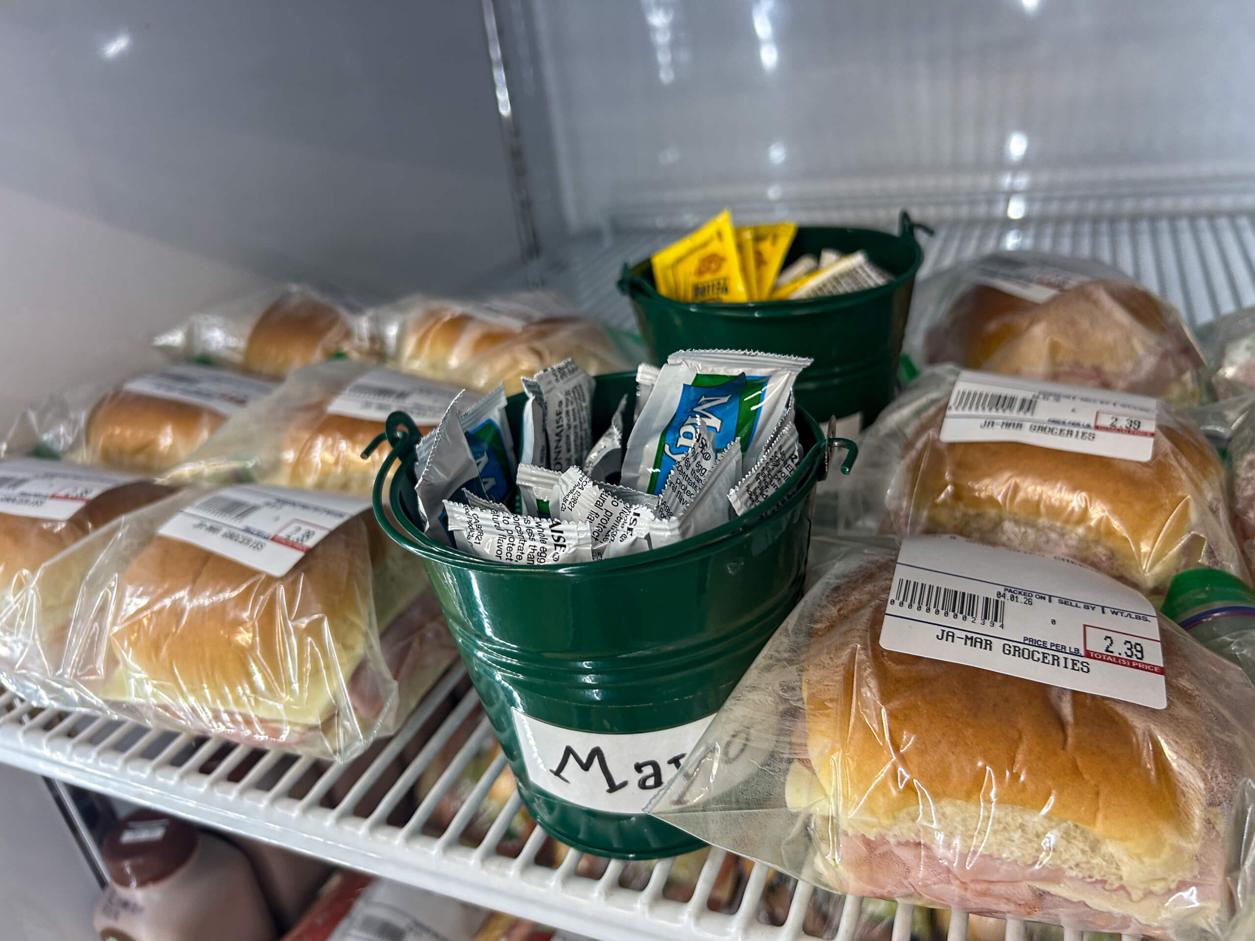 Jamar Groceries Kutztown PA Pre-packaged sandwiches and small green buckets containing mayonnaise and mustard packets are displayed on a refrigerator shelf.