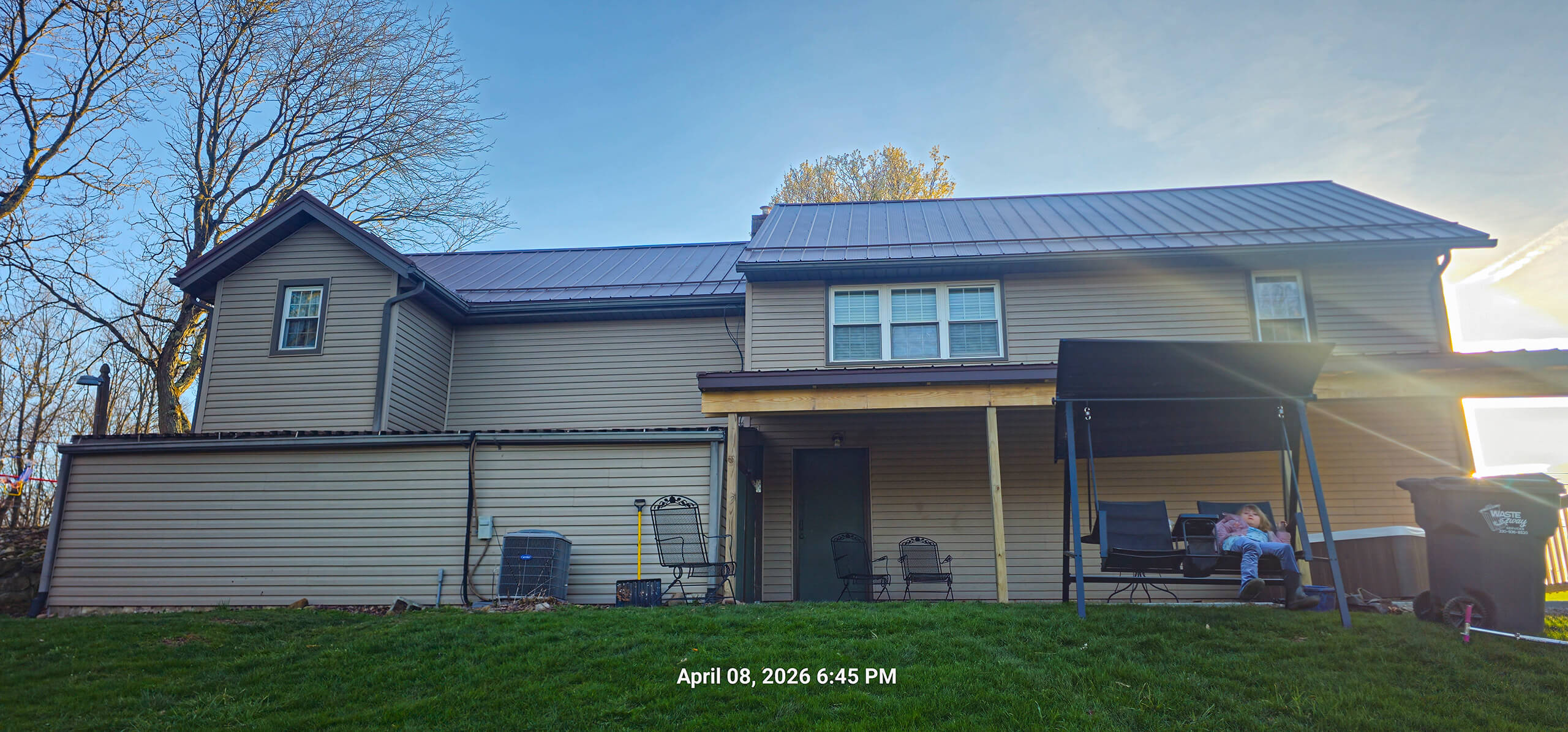 Nelson Weaver Metal Roofing Carrollton OH A two-story beige house with a metal roof, a covered patio, outdoor chairs, and a swing. The yard is grassy, and the time stamp reads April 08, 2026, 6:45 PM.