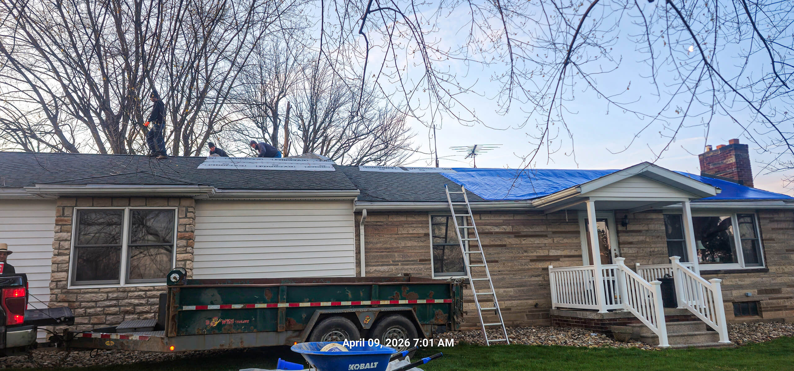 Nelson Weaver Metal Roofing Carrollton OH Workers repair a house roof, with part of the roof covered by a blue tarp; a ladder and trailer are positioned outside, and trees are visible in the background.