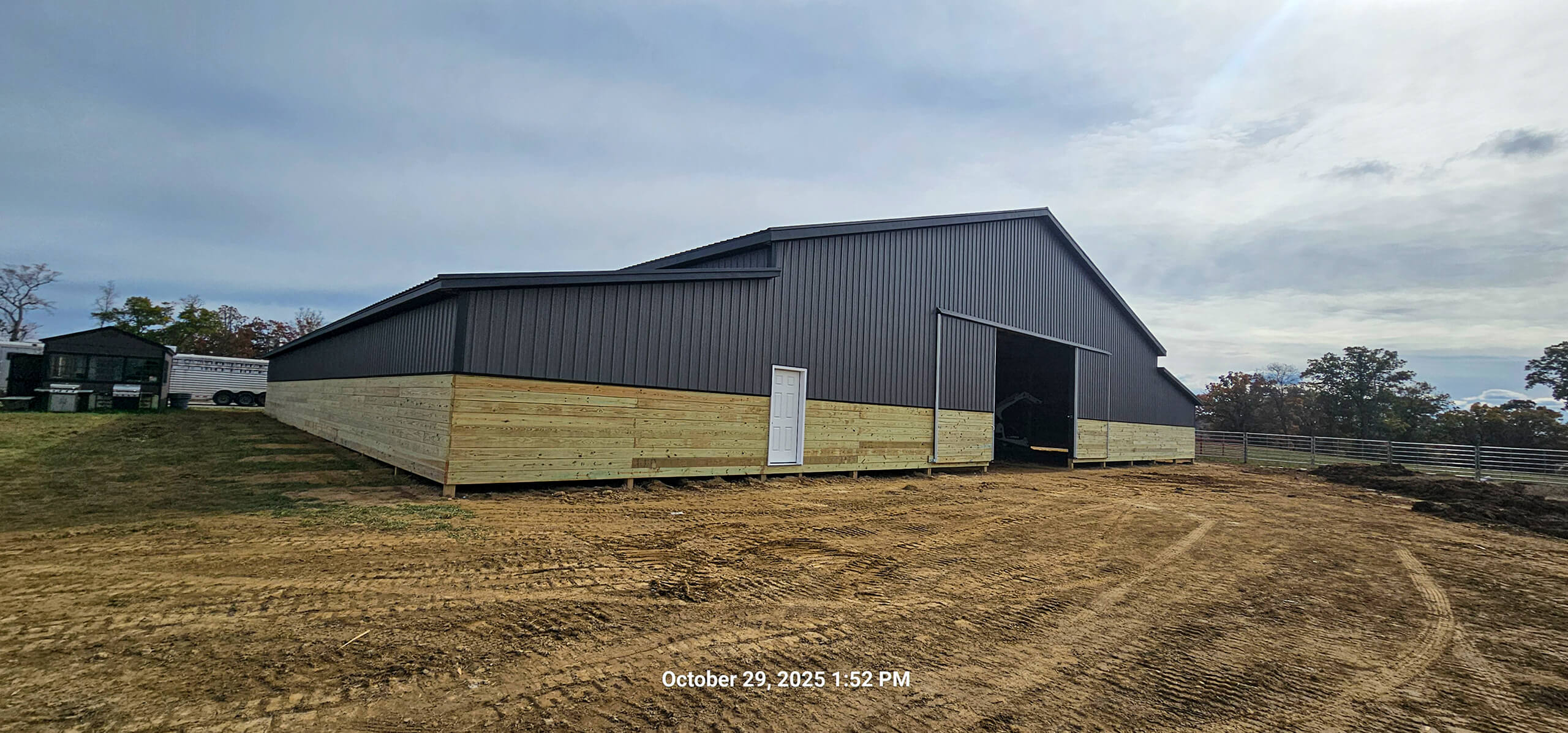 Nelson Weaver Metal Roofing Carrollton OH Large barn with dark metal siding and light wooden lower panels stands on bare ground under an overcast sky. A white door is visible on the side. Date and time stamped at bottom.