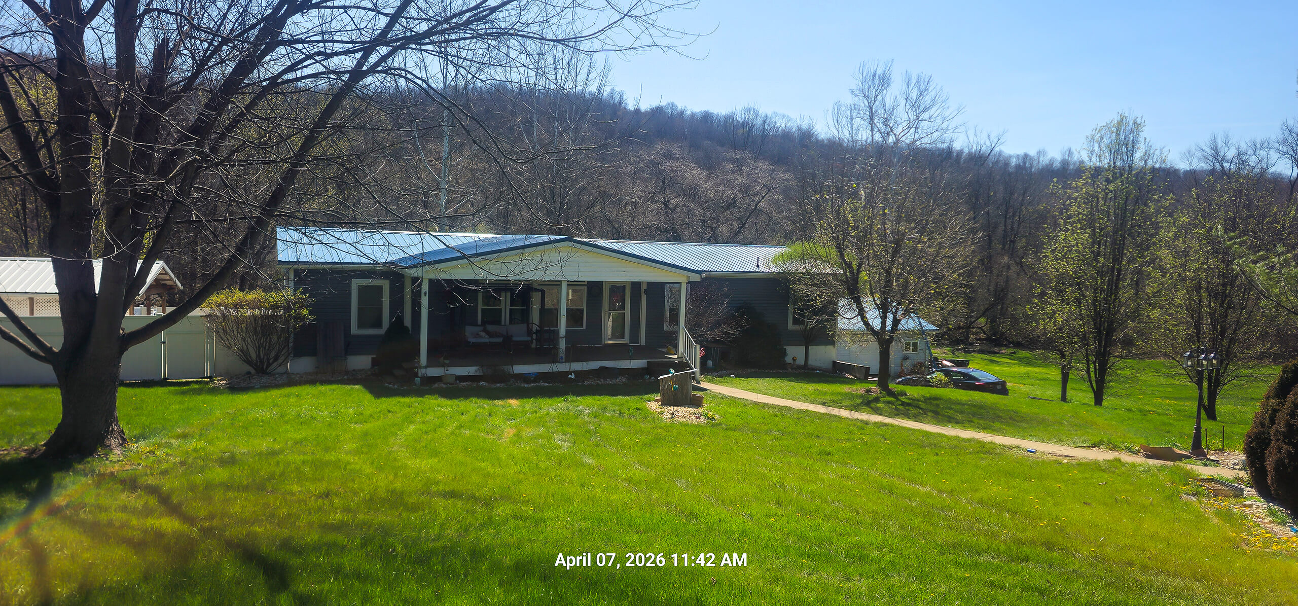 Nelson Weaver Metal Roofing Carrollton OH A single-story house with a covered porch sits on a grassy lawn surrounded by leafless and budding trees. Hills are visible in the background. The date April 07, 2026, is displayed.