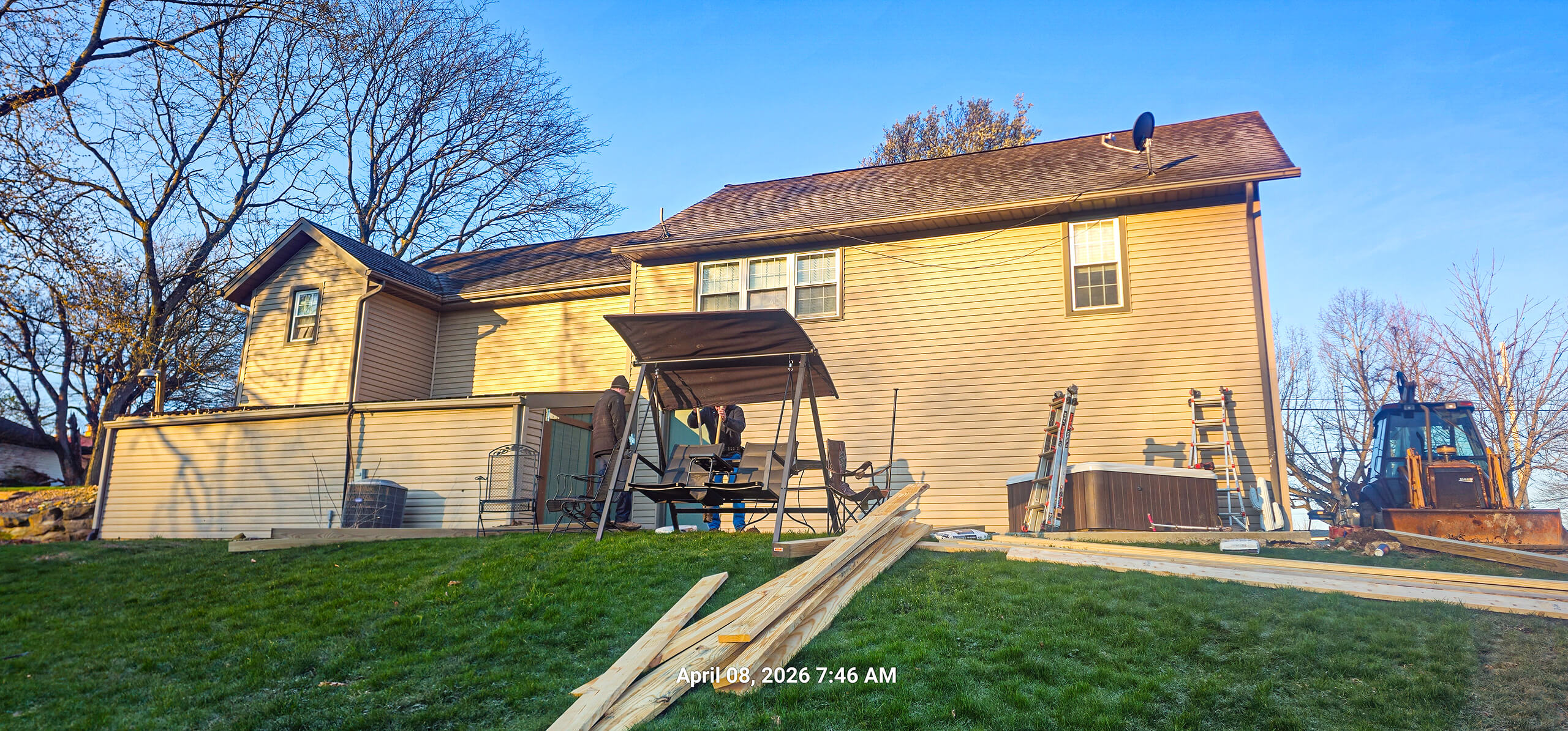 Nelson Weaver Metal Roofing Carrollton OH Backyard of a beige house with construction materials on the grass, a swing set, a ladder, and a yellow excavator in the background. Photo taken on April 8, 2026 at 7:46 AM.