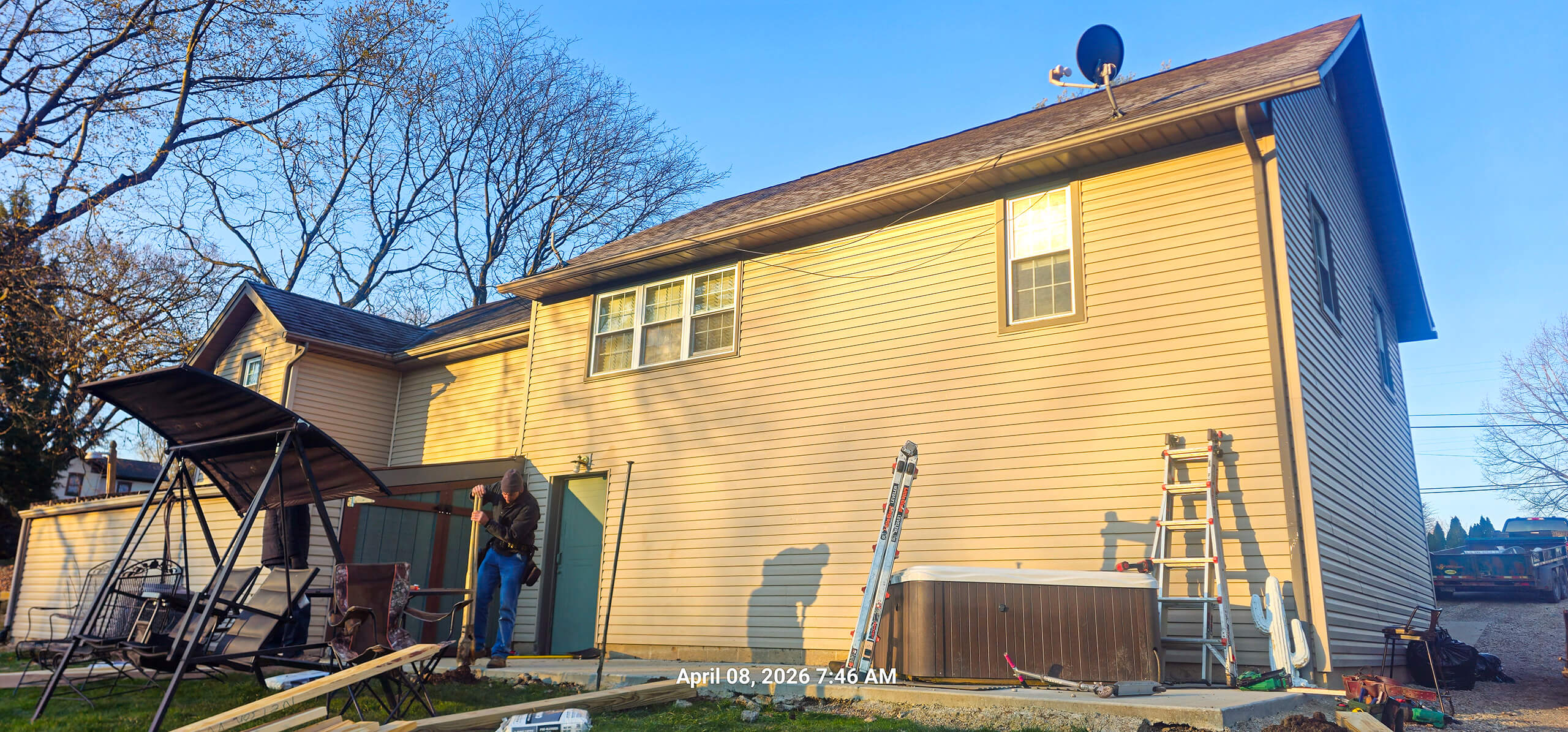 Nelson Weaver Metal Roofing Carrollton OH A person stands near the back door of a beige two-story house with outdoor furniture, ladders, and a satellite dish visible; timestamp reads April 08, 2026, 7:46 AM.