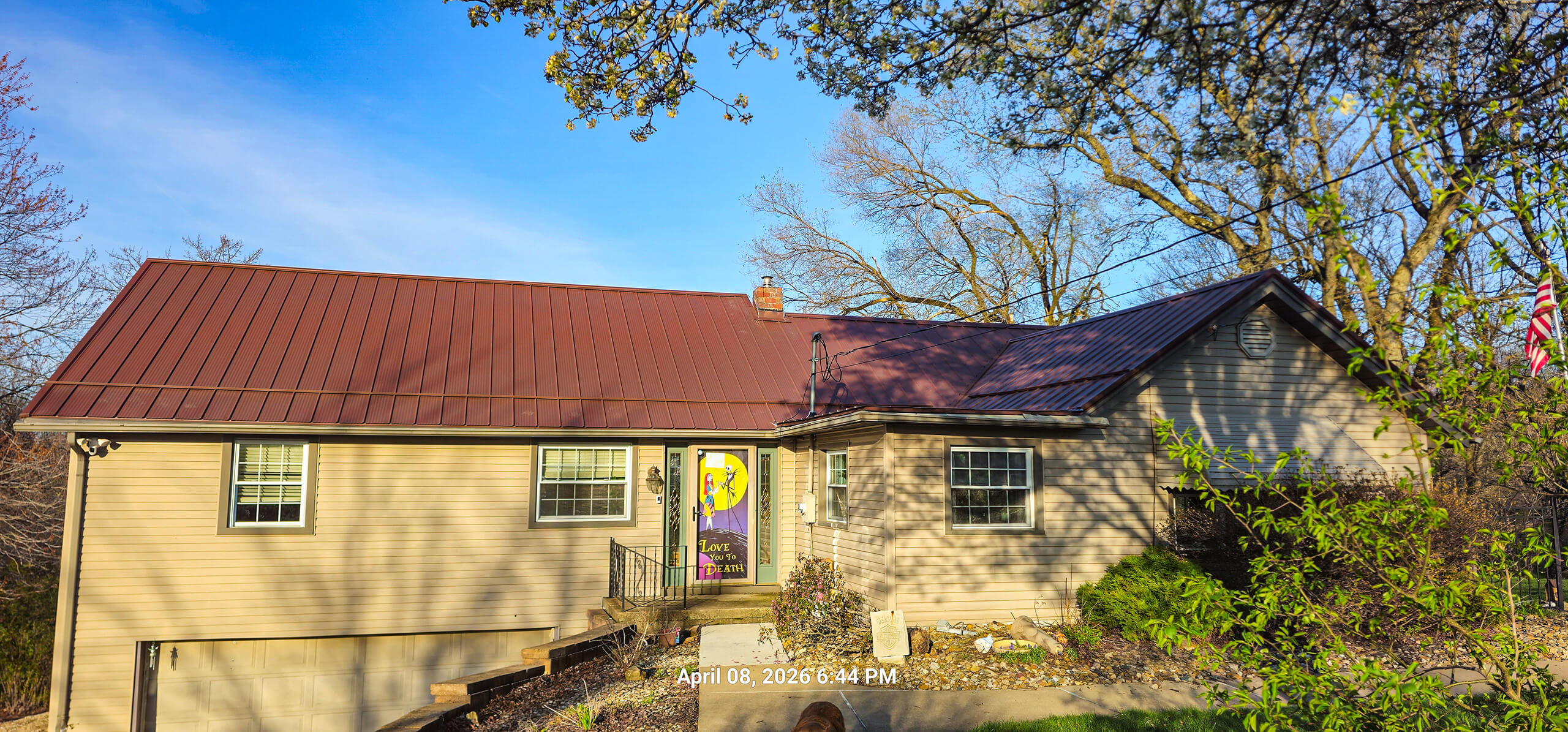 Nelson Weaver Metal Roofing Carrollton OH Single-story beige house with a red metal roof, front steps, a colorful flag by the door, surrounded by trees and shrubs. Time stamp reads "April 08, 2023 6:44 PM.