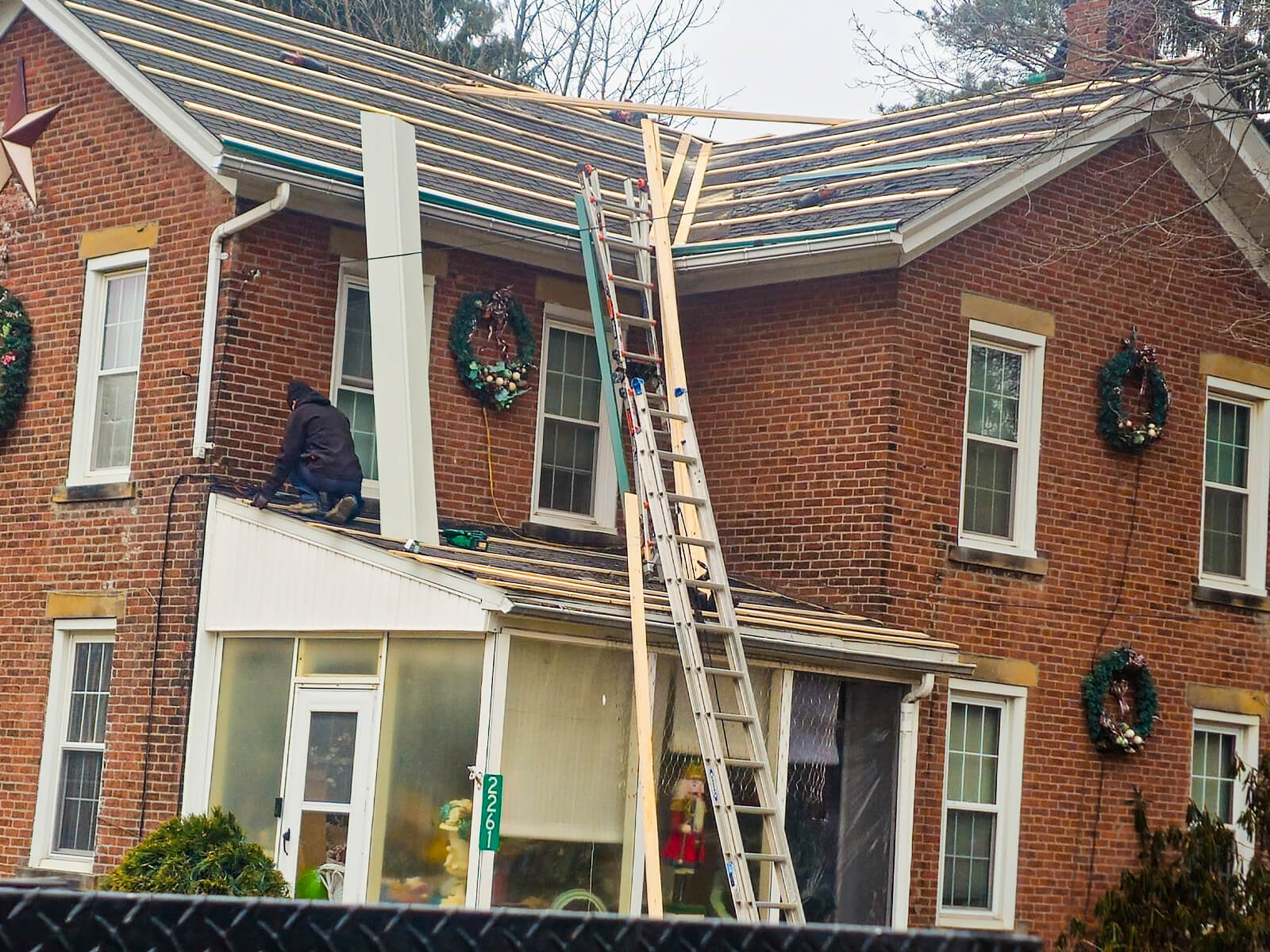 Nelson Weaver Metal Roofing Carrollton OH A person works on the roof of a brick house under renovation, with ladders and roofing materials visible and holiday wreaths on the windows.