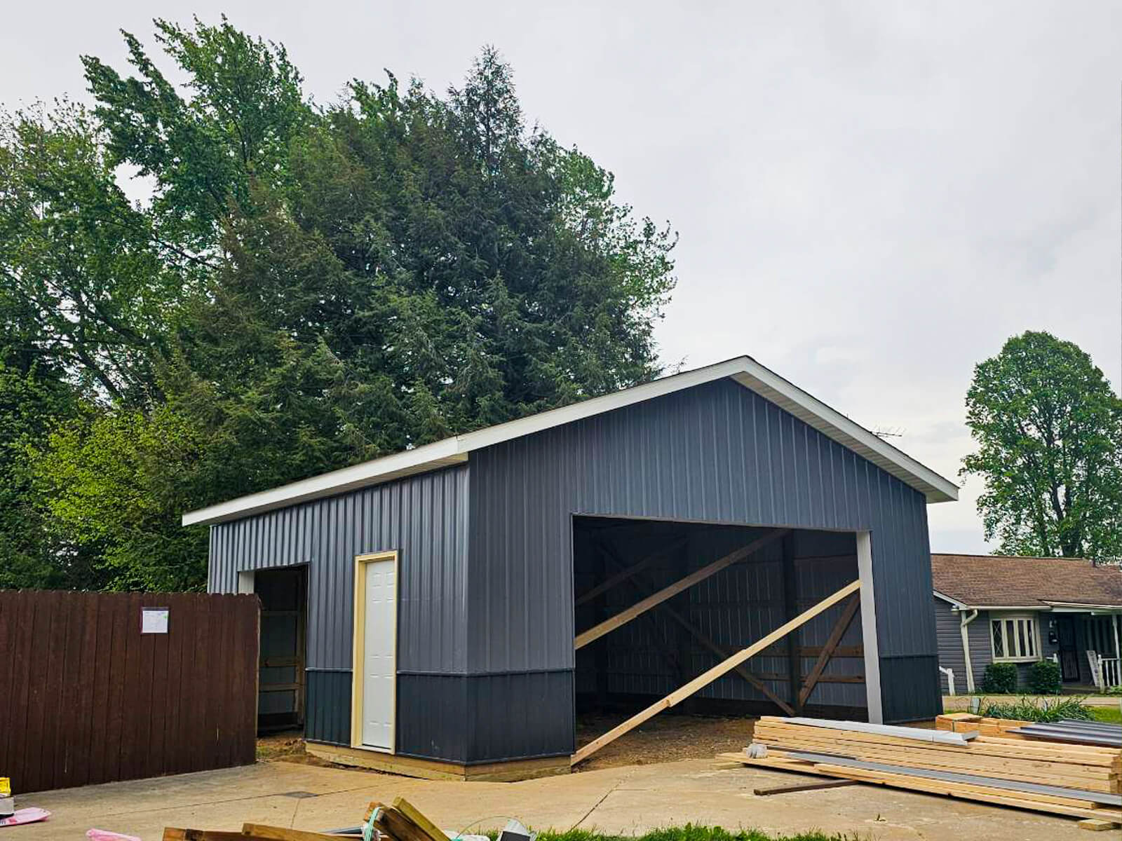 Nelson Weaver Metal Roofing Carrollton OH A partially constructed gray metal garage with open front, white door, and wood supports, situated next to a house and trees on a cloudy day.