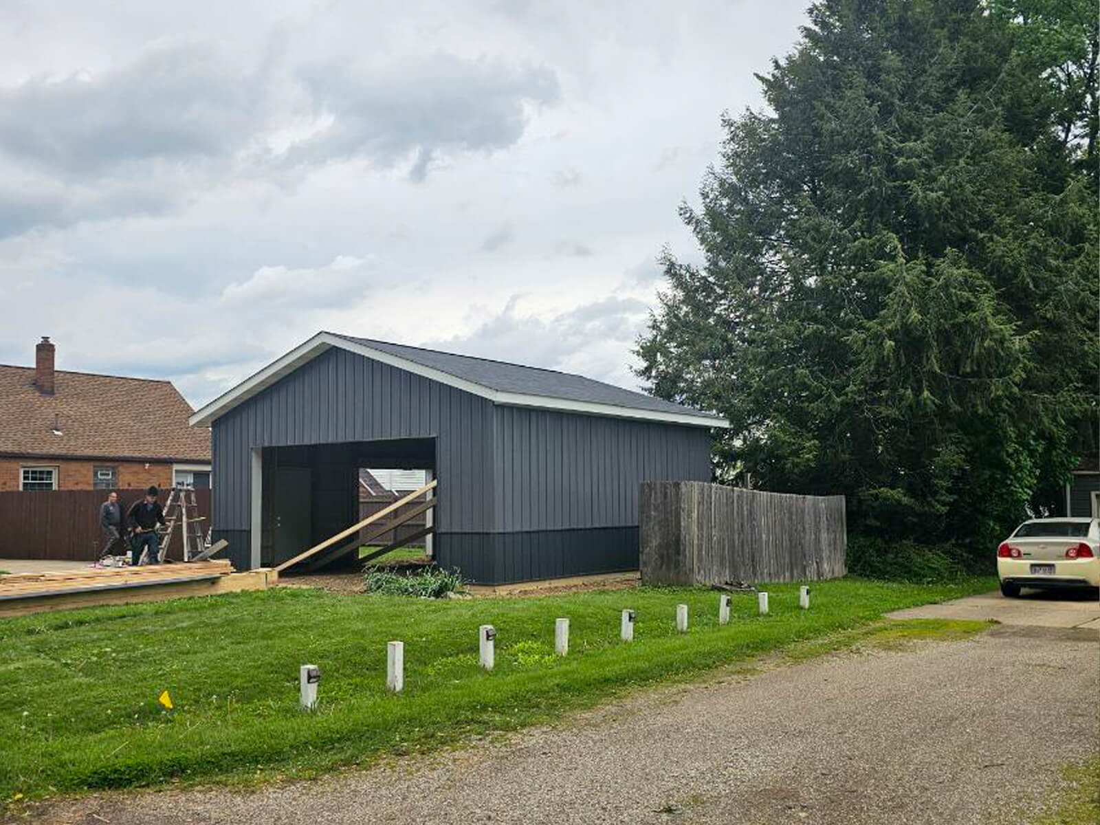 Nelson Weaver Metal Roofing Carrollton OH A gray garage under construction with workers on the left, adjacent to a wooden fence, trees, and a parked white car on the right.