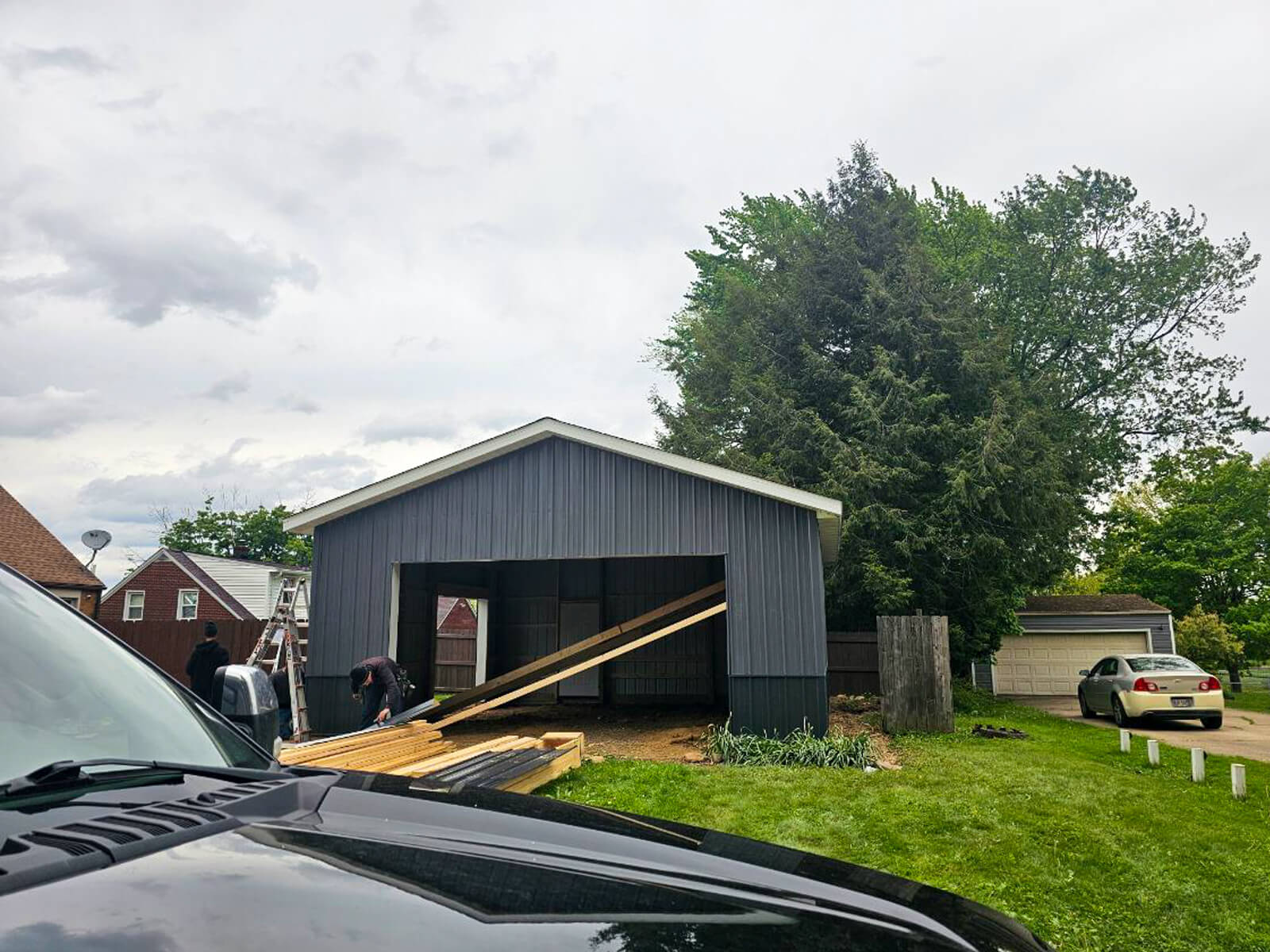 Nelson Weaver Metal Roofing Carrollton OHa A partially constructed garage or shed with dark paneling, a pile of lumber, and a person working beside it; cars and houses are visible in the background.