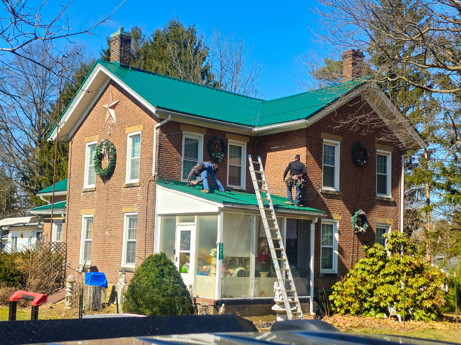 Nelson Weaver Metal Roofing Carrollton OH Two people are installing holiday wreaths on the windows and roof of a two-story brick house using ladders on a clear, sunny day.