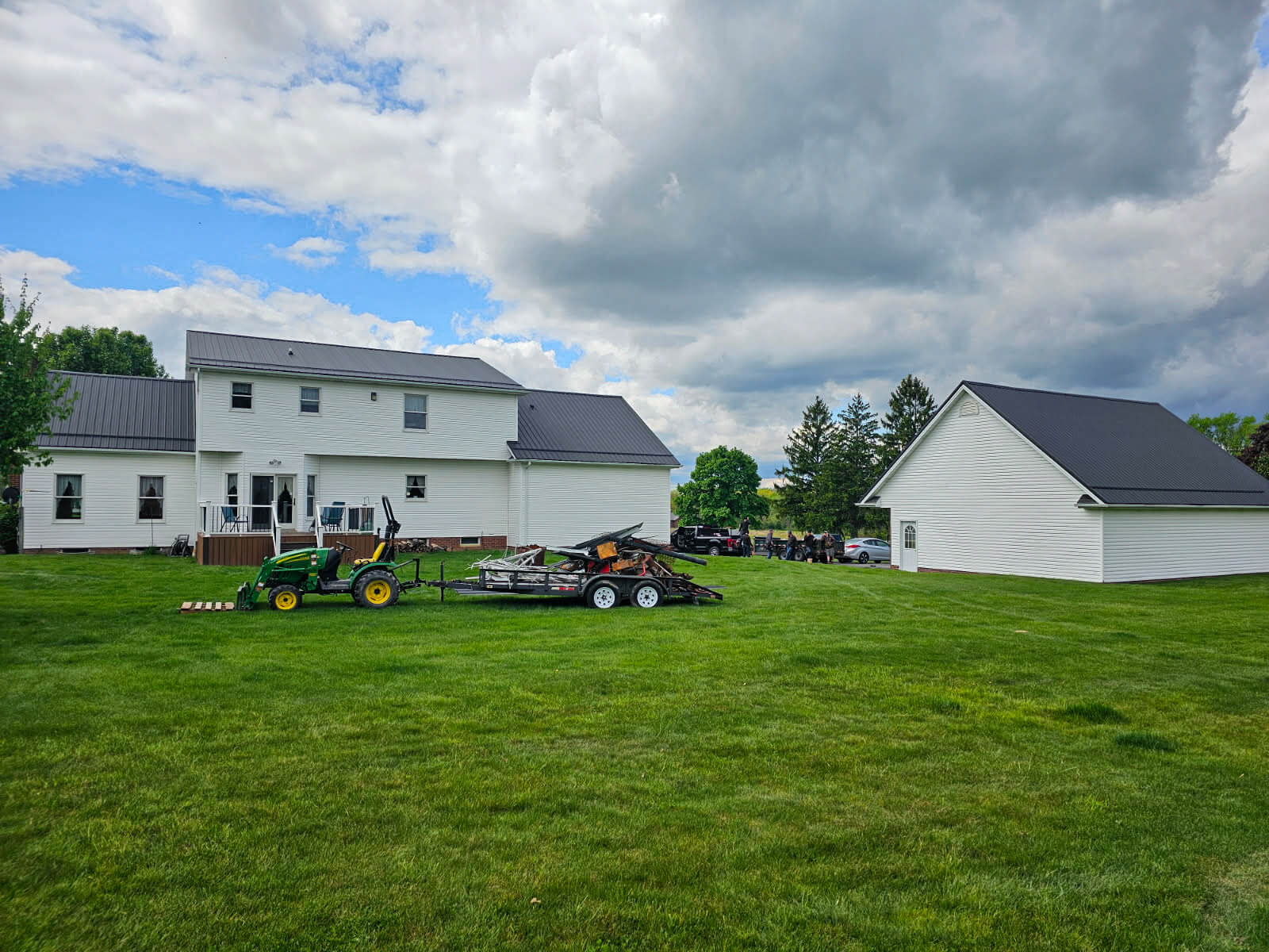 Nelson Weaver Metal Roofing Carrollton OH A white two-story house and detached garage stand on a large grassy lawn, with a green tractor and trailer parked nearby under a cloudy sky.