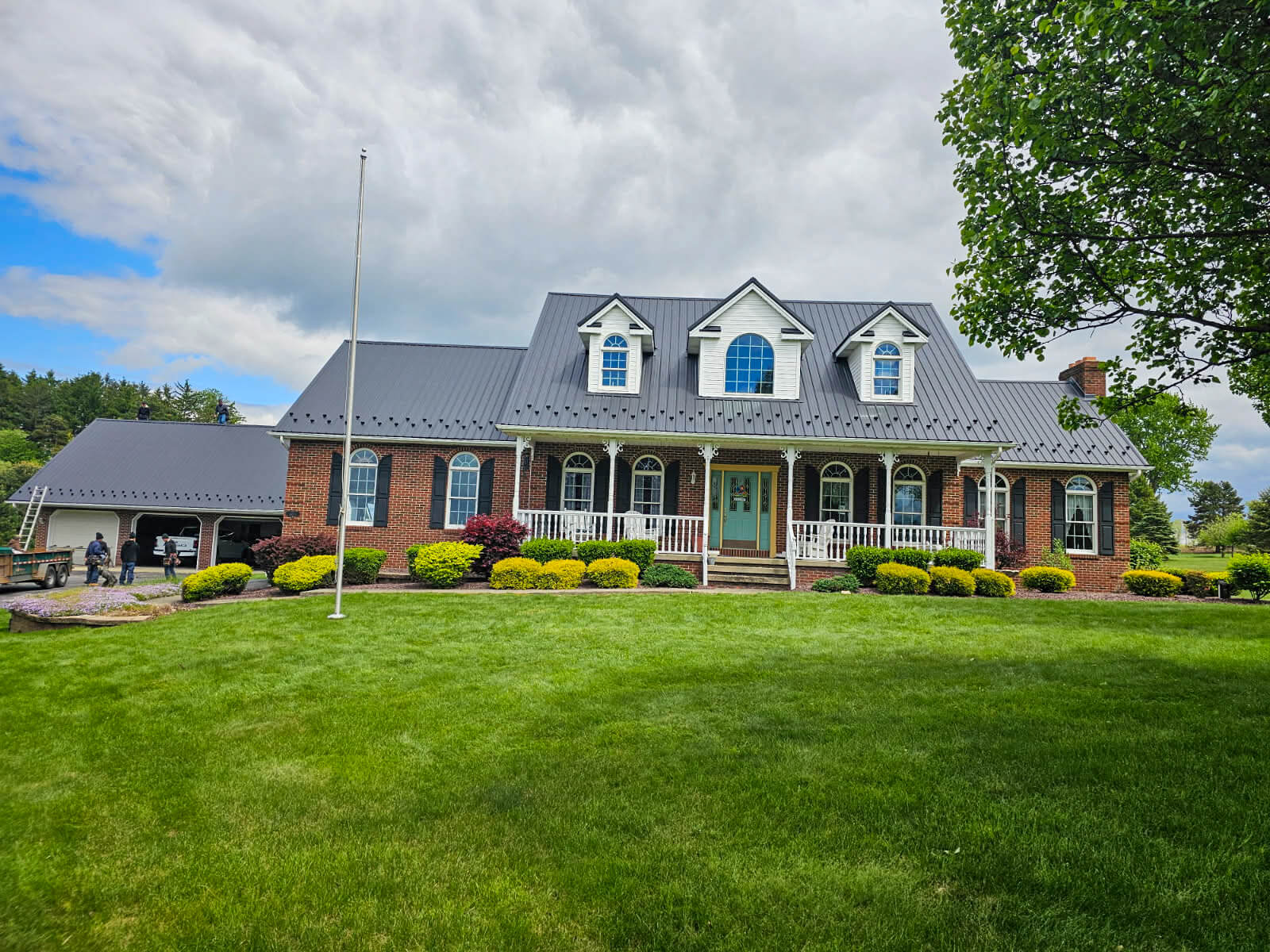 Nelson Weaver Metal Roofing Carrollton OH A large brick house with a gray metal roof, white trim, and dormer windows, surrounded by a manicured lawn and shrubs under a partly cloudy sky.