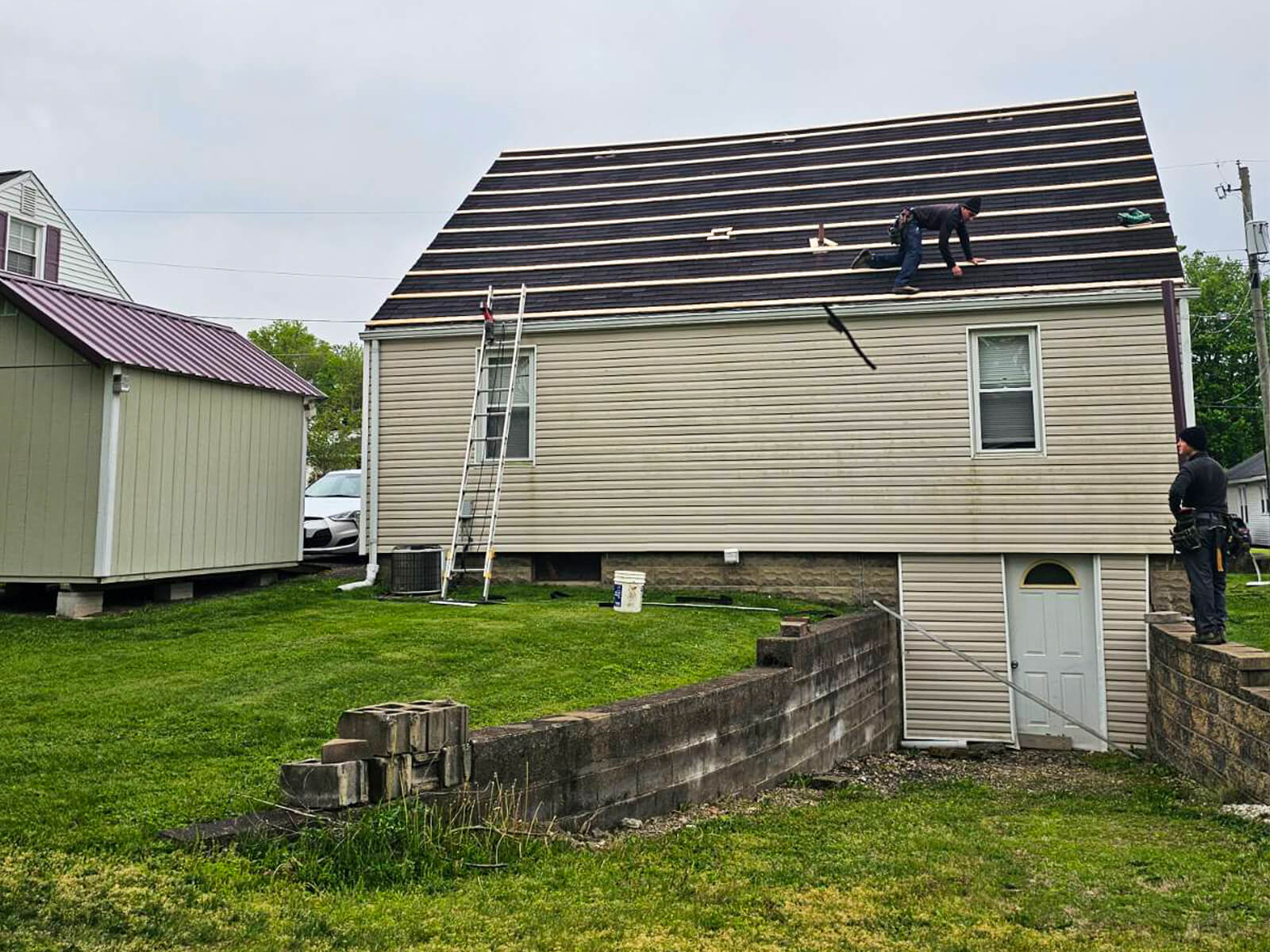 Nelson Weaver Metal Roofing Carrollton OH Two workers are installing roofing materials on a beige house, with ladders and tools visible on the ground and roof.