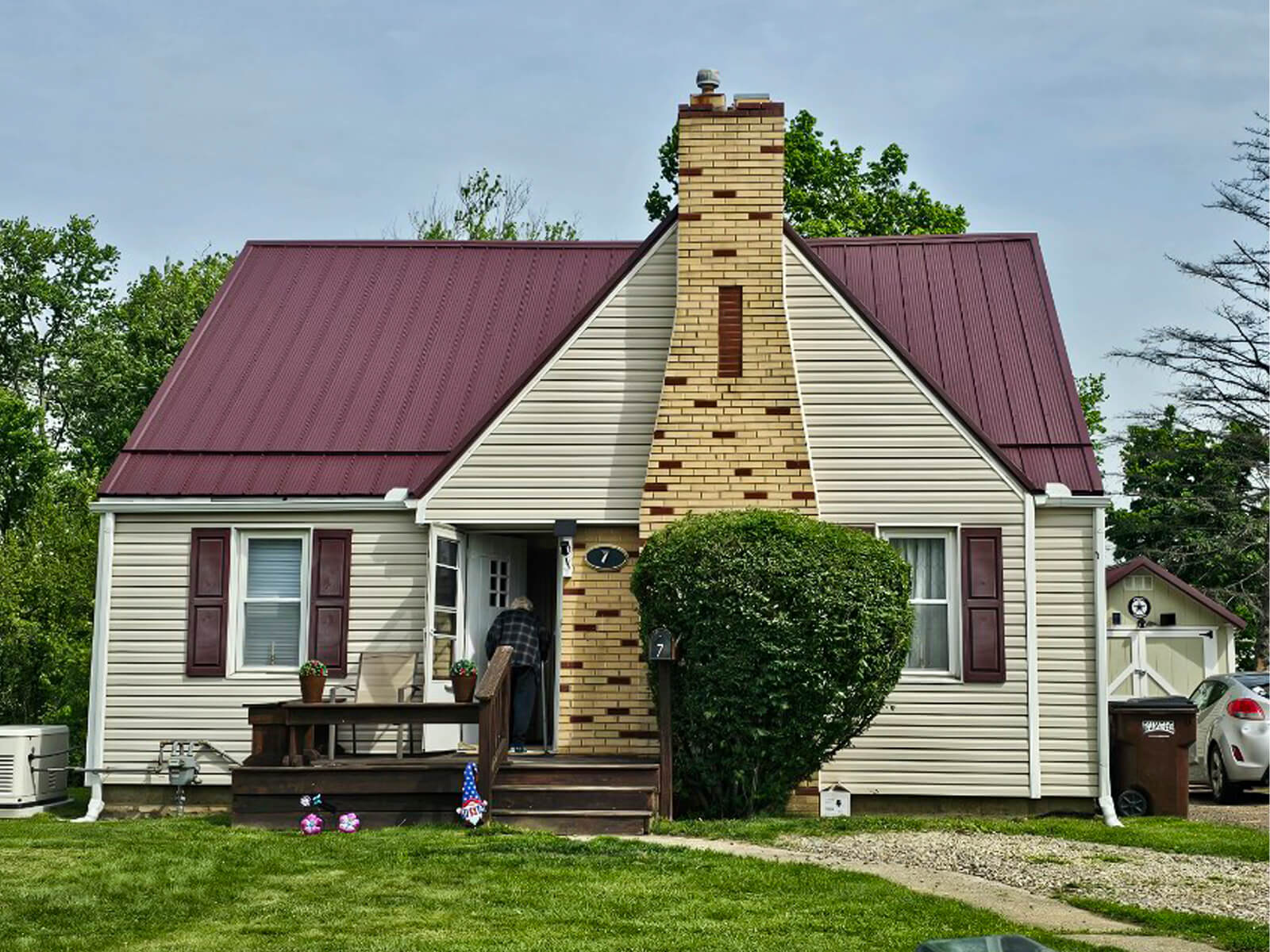 Nelson Weaver Metal Roofing Carrollton OH A small beige house with a steep maroon metal roof, a brick chimney, front porch, shrubs, and a driveway with a white car parked to the right.