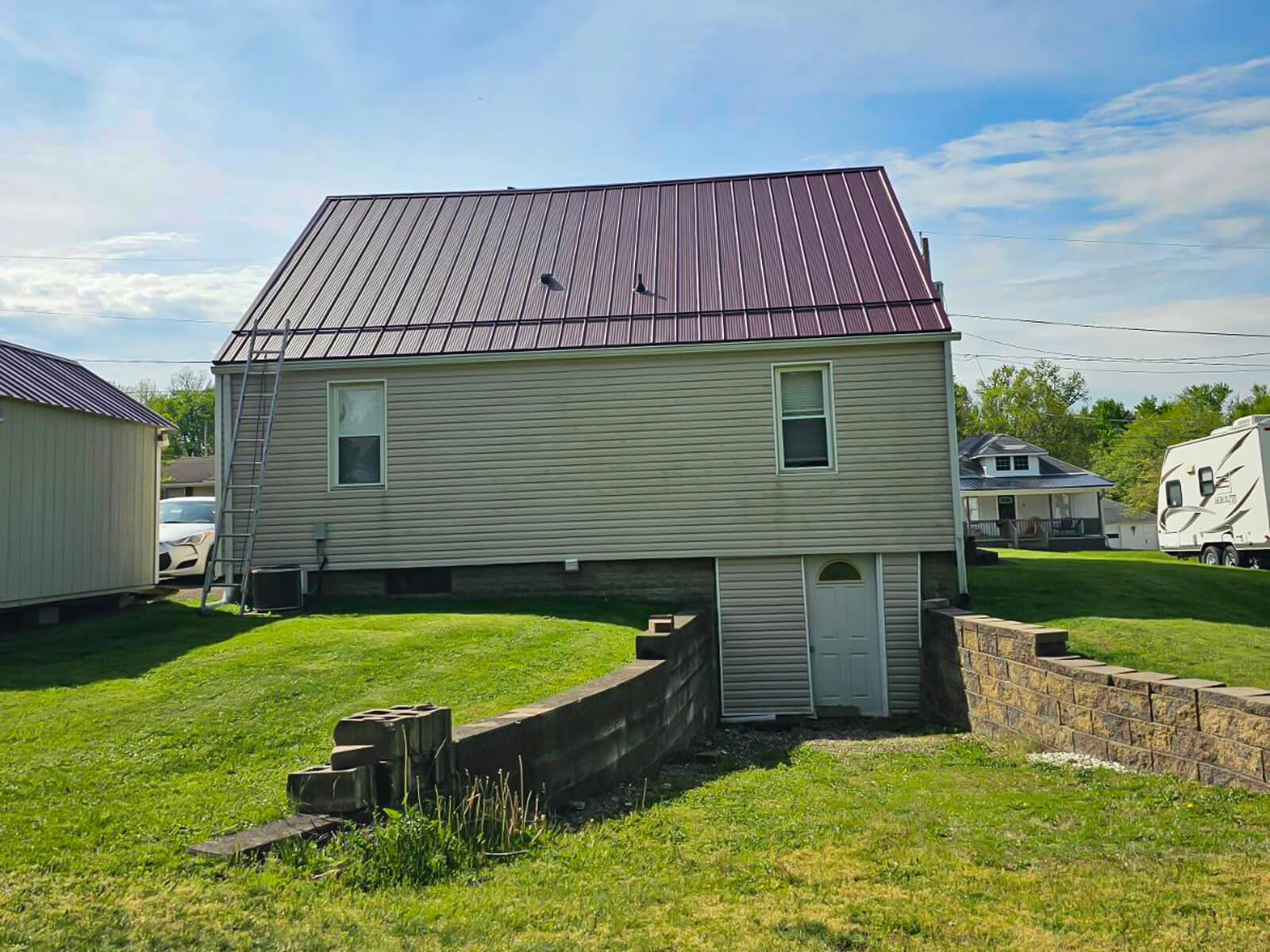 Nelson Weaver Metal Roofing Carrollton OH A beige house with a red metal roof, basement door, and concrete retaining walls on a grassy lawn under a partly cloudy sky.