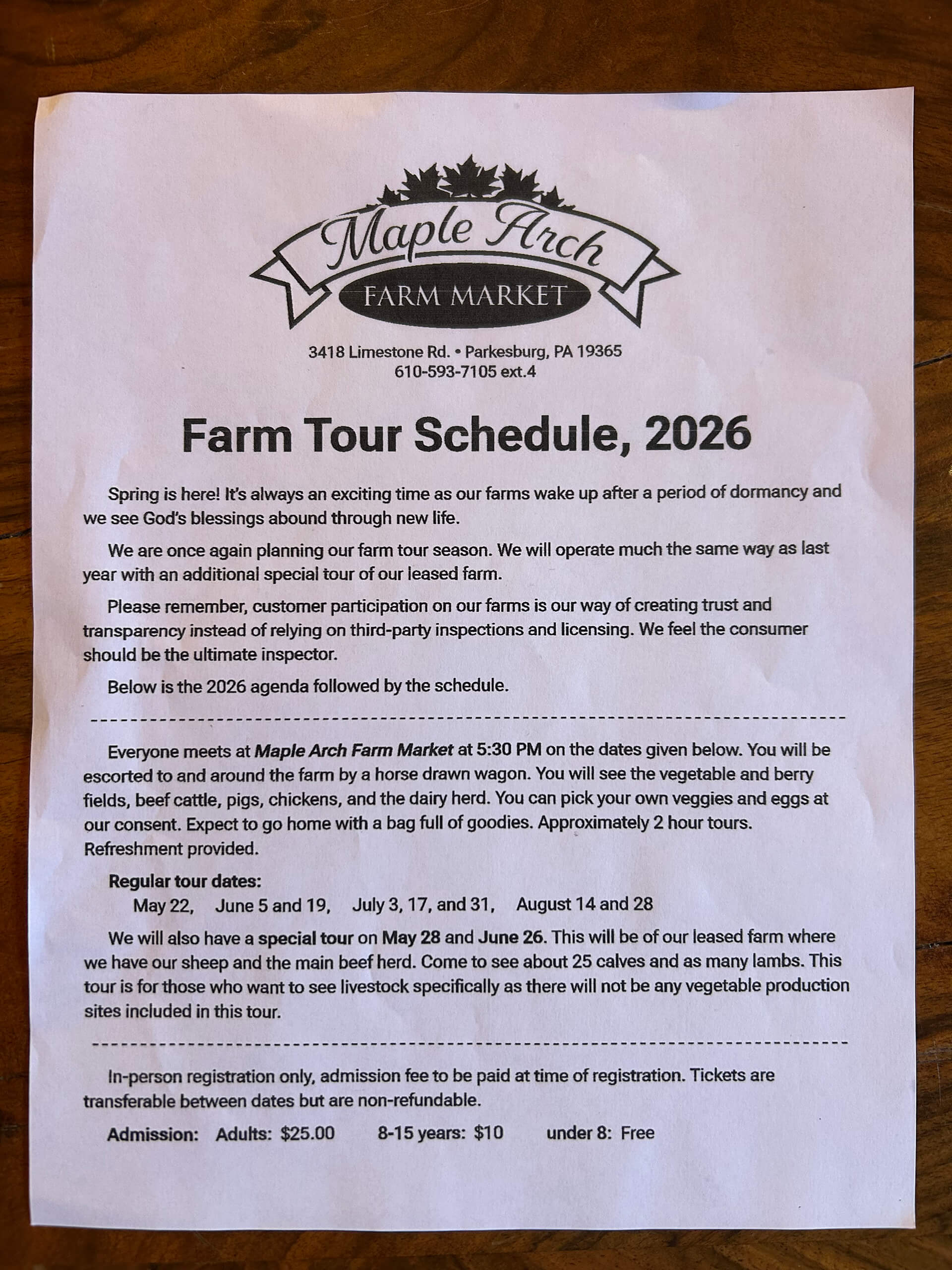 5.5.26 Maple Arch Farm Market Ad 2 A printed flyer for Maple Arch Farm Market displays the 2026 farm tour schedule, dates, times, admission prices, and tour details on a wooden surface.