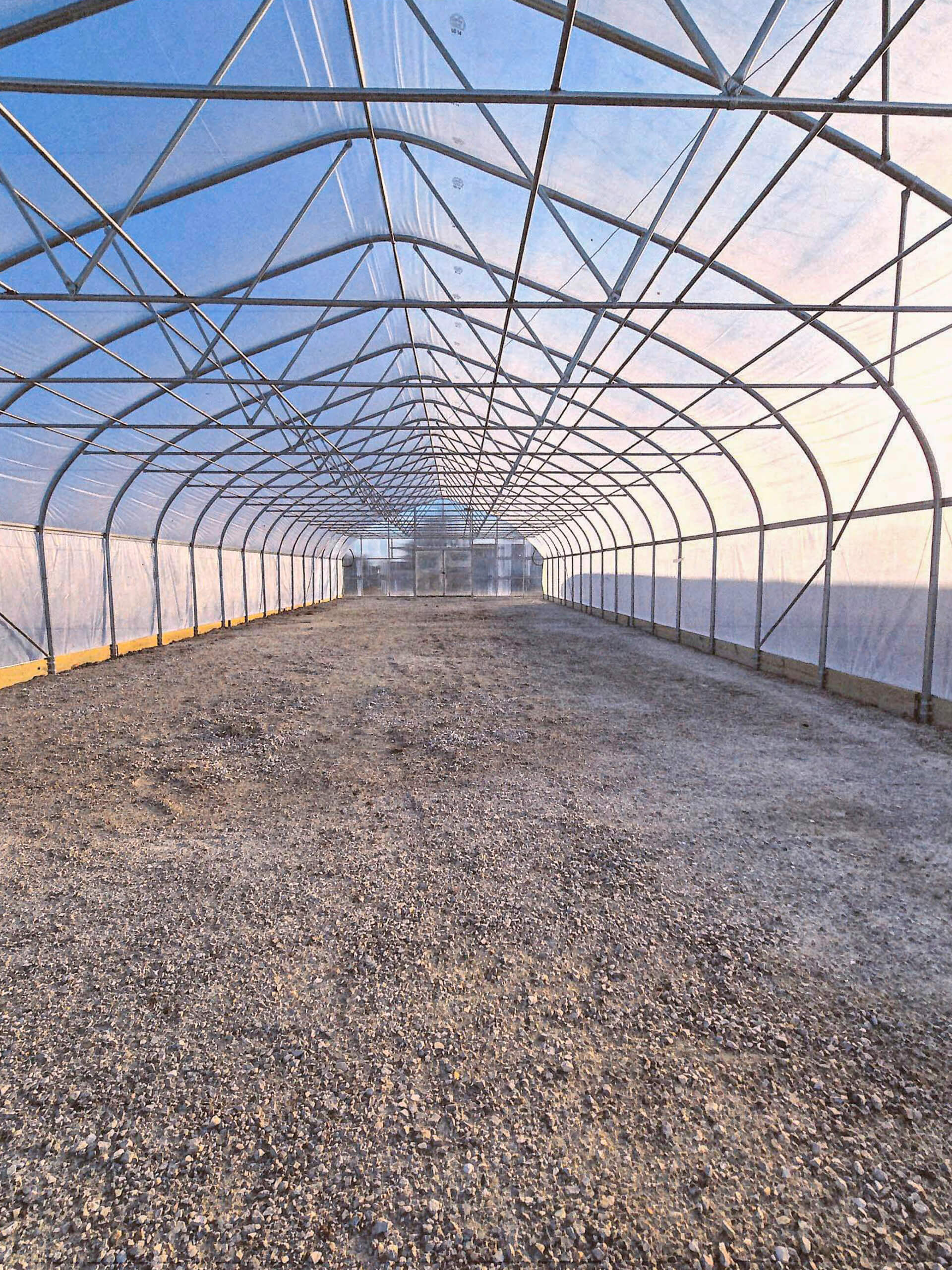 Sunny Crest Acres Versailles MO Interior of an empty greenhouse with a curved metal frame, transparent plastic covering, and a gravel floor.