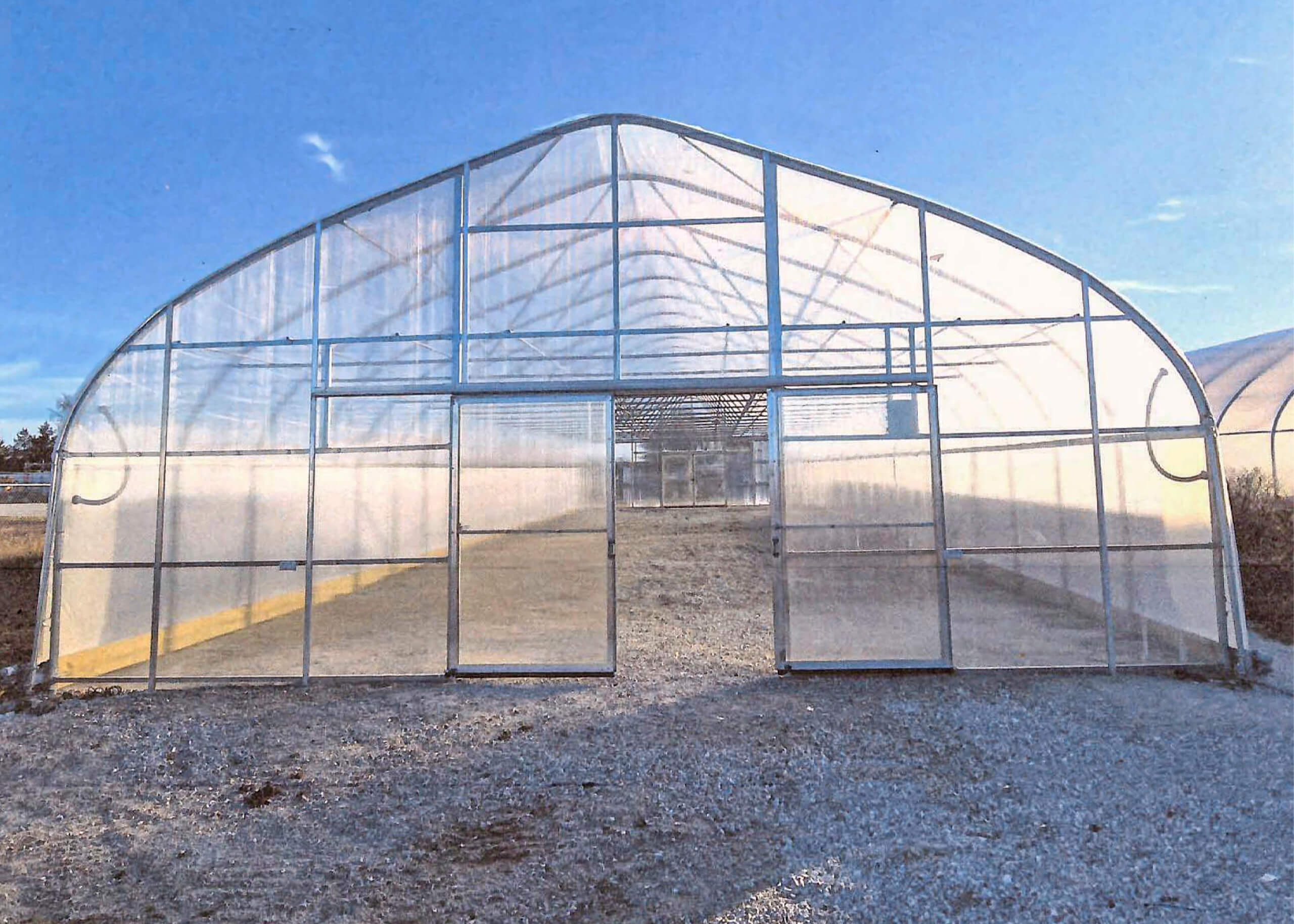 5.23.26 SunSunny Crest Acres Versailles MOny Crest Acres Gallery 2 A clear plastic greenhouse with a peaked roof and open doorway stands on a gravel surface under a blue sky.