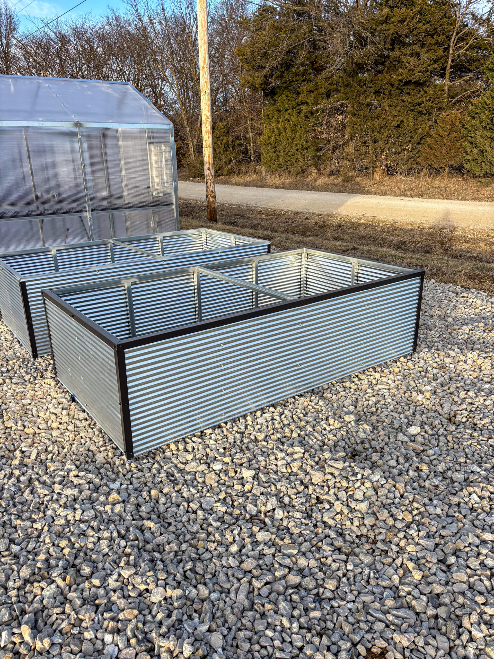Sunny Crest Acres Versailles MO A metal raised garden bed frame sits on gravel beside a greenhouse, with trees and a dirt road in the background.
