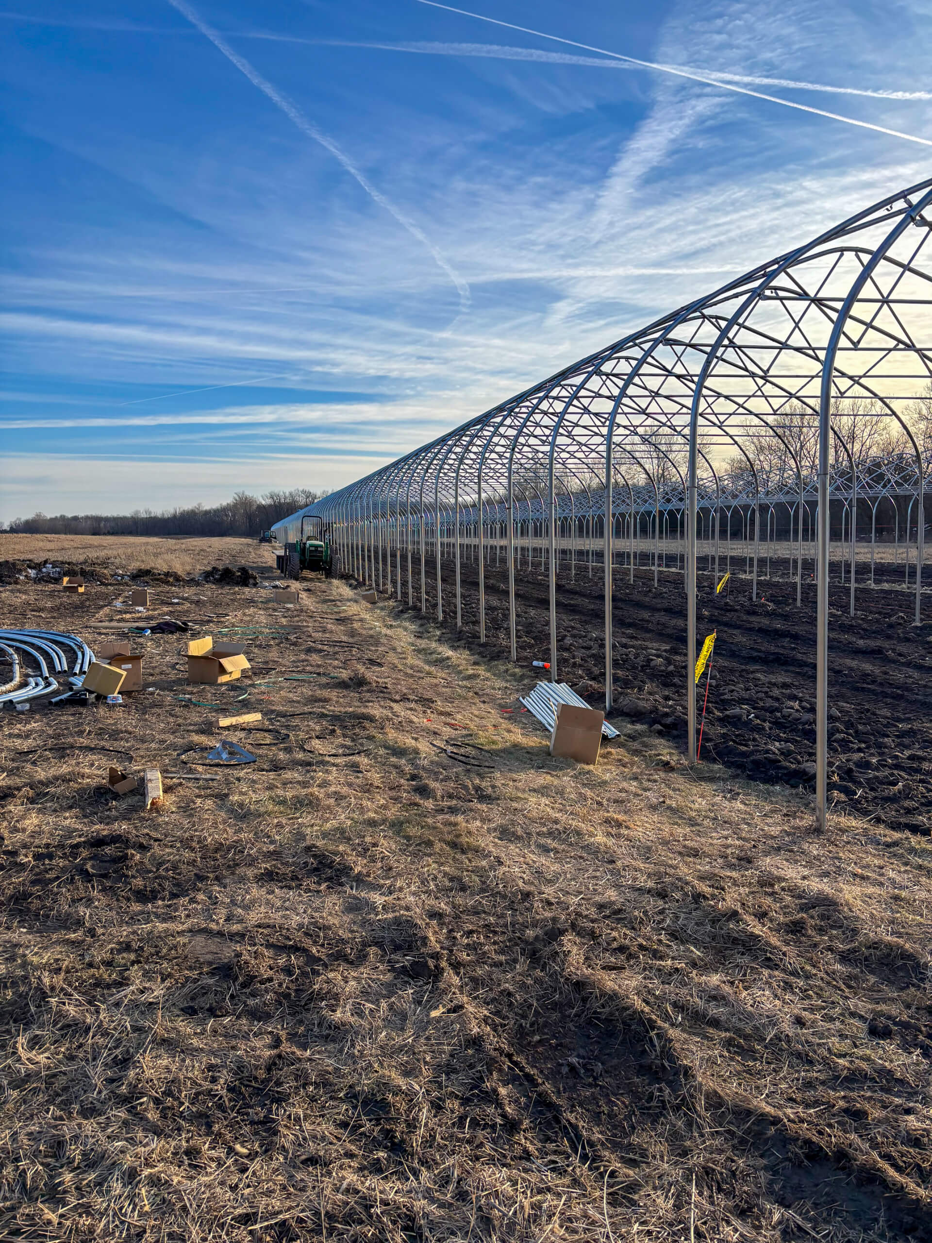Sunny Crest Acres Versailles MO A metal greenhouse frame stands on a field under a blue sky, with construction materials and equipment scattered on the ground nearby.