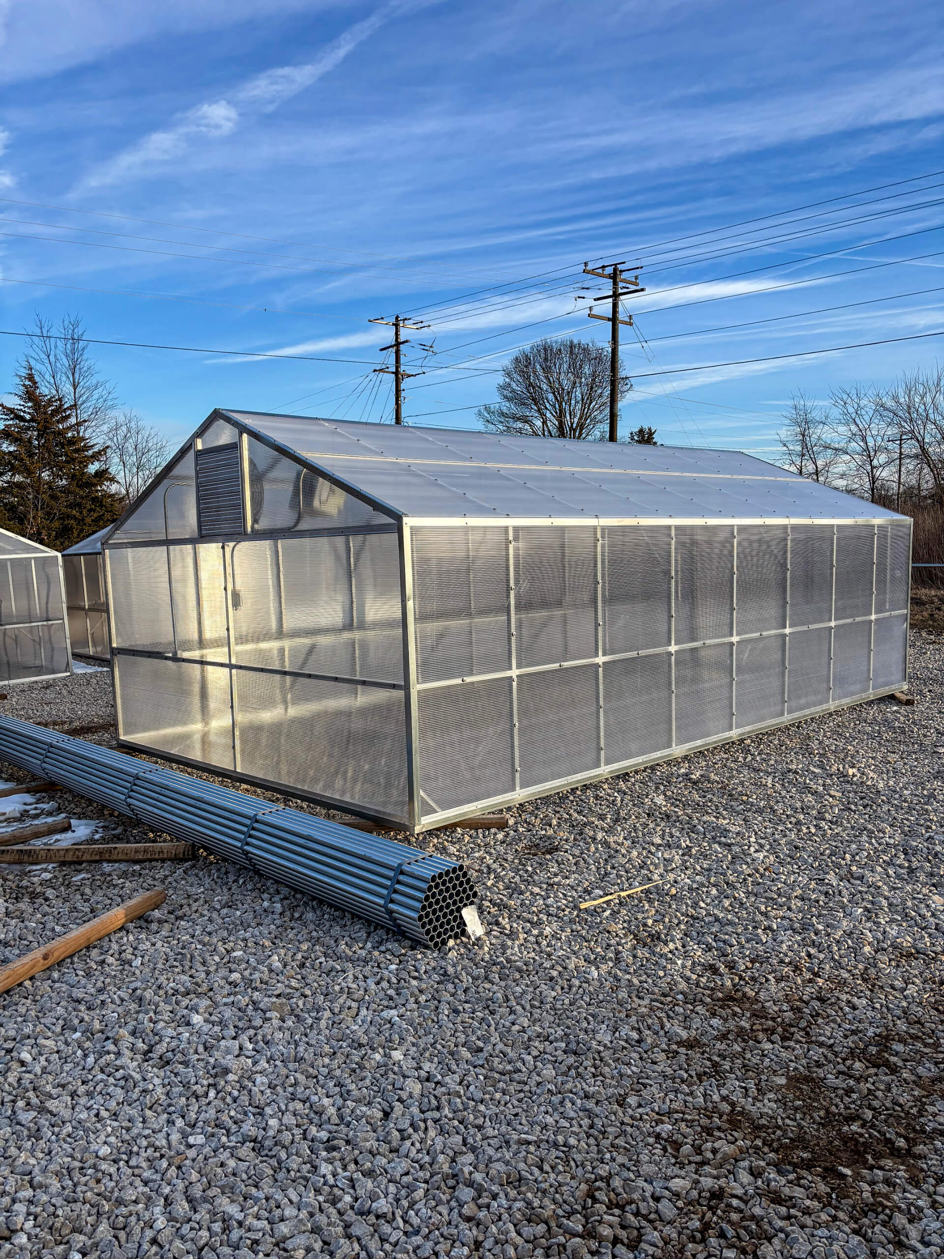 Sunny Crest Acres Versailles MO A metal-framed greenhouse with clear panels stands on a gravel lot under a blue sky, with metal pipes lying on the ground nearby.
