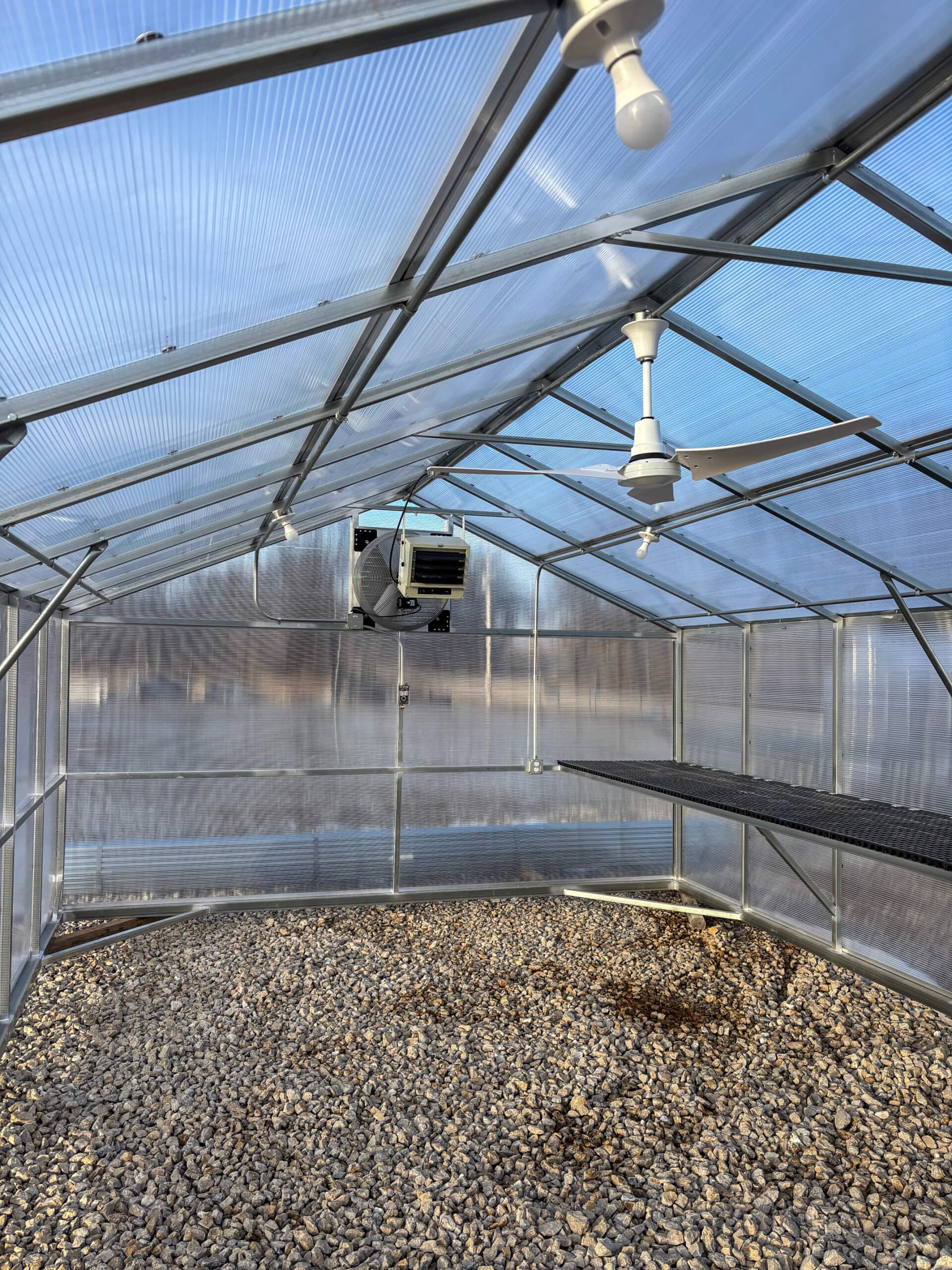 Sunny Crest Acres Versailles MO Interior of an empty greenhouse with transparent walls, gravel floor, a black metal bench, ceiling fans, and a mounted heater.