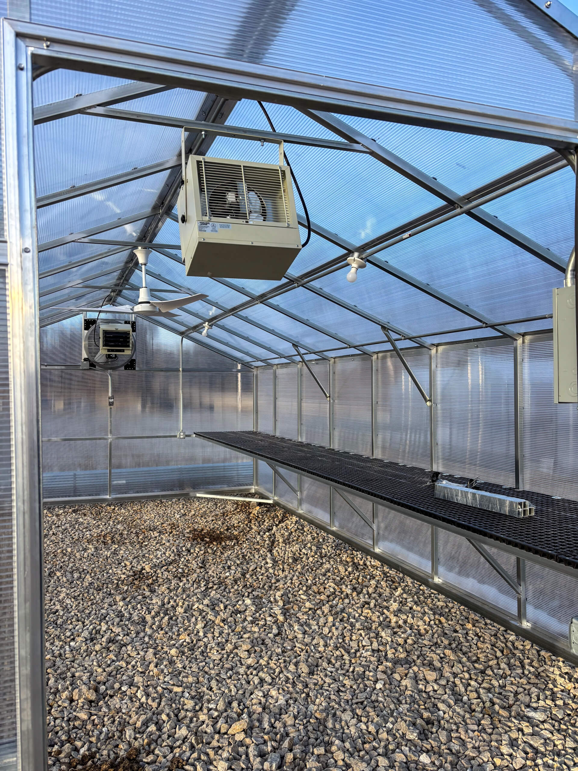Sunny Crest Acres Versailles MO Interior of an empty greenhouse with two ceiling-mounted heaters, gravel floor, and metal shelving along one side.