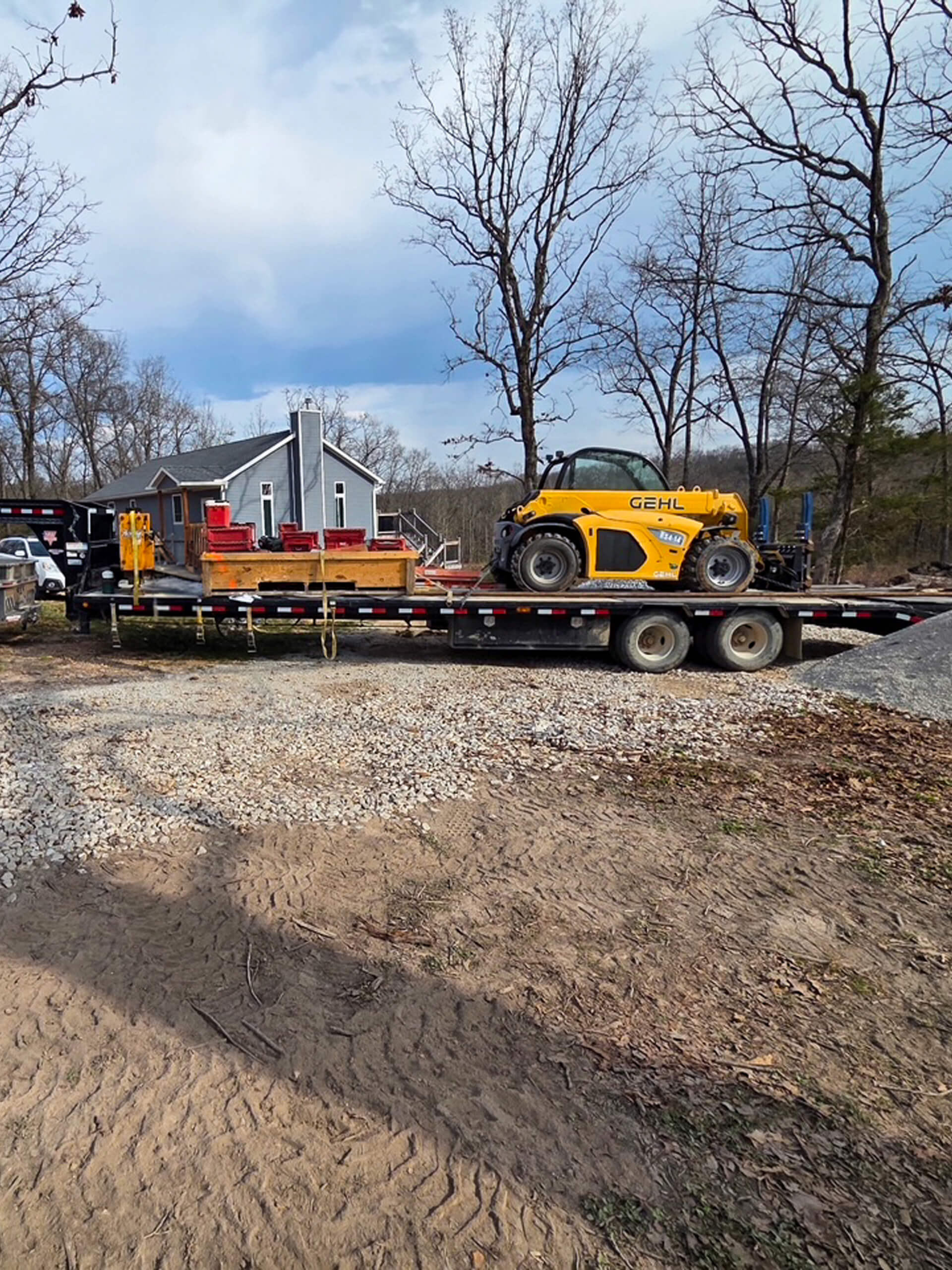 Sunny Crest Acres Versailles MO A flatbed trailer loaded with a yellow Gehl telehandler and other construction materials is parked on a gravel driveway near a house and trees.