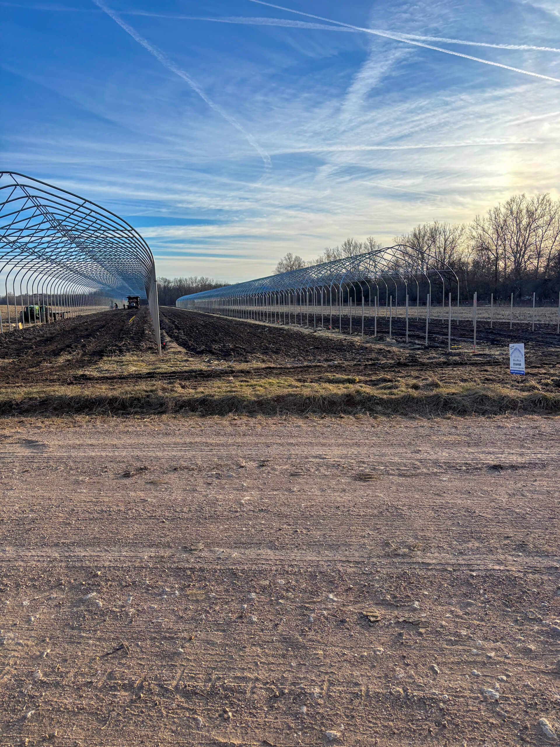 Sunny Crest Acres Versailles MO Metal frames for greenhouse structures are set up on a muddy field under a clear sky, with a dirt road in the foreground and trees in the background.