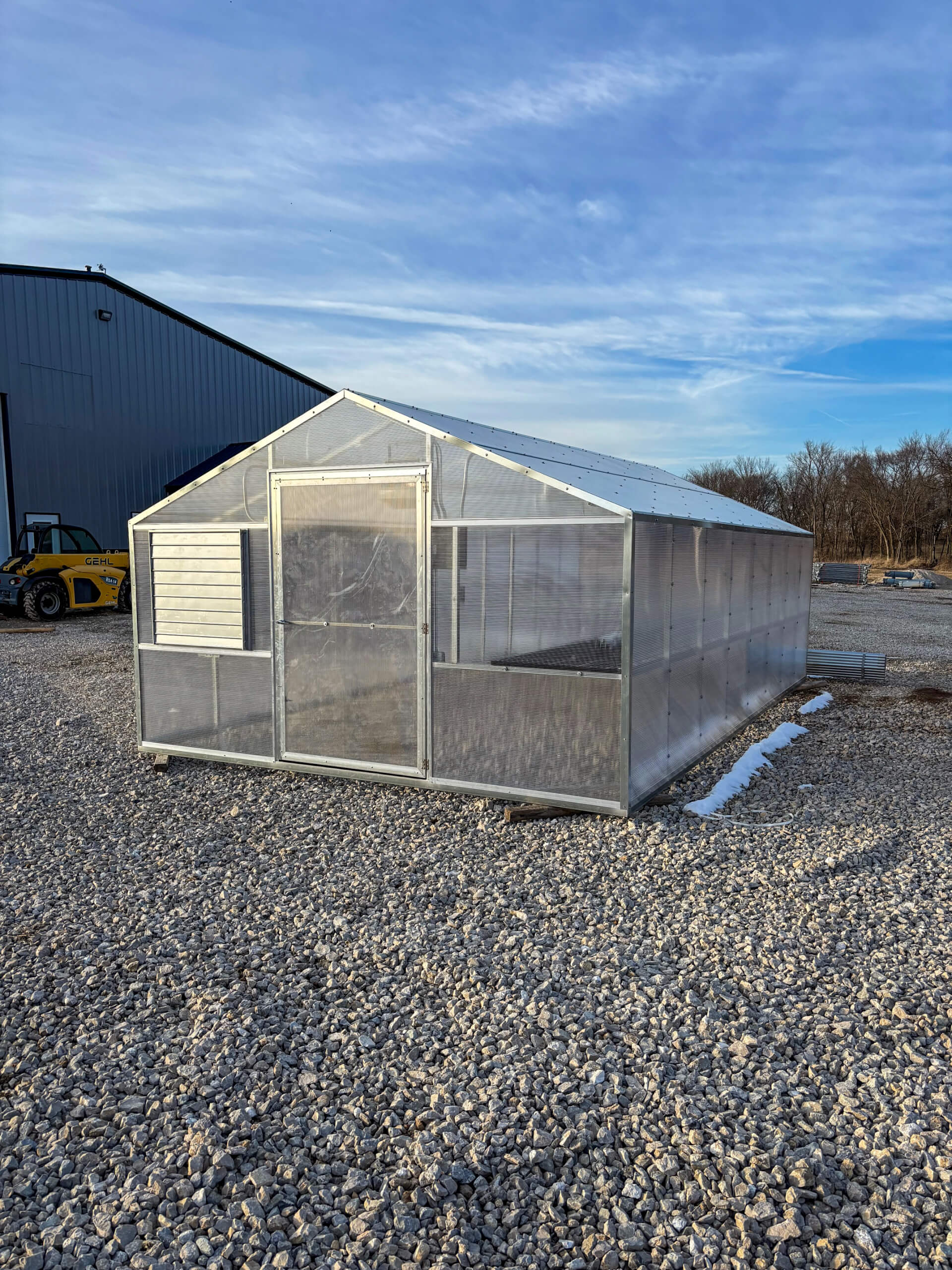 Sunny Crest Acres Versailles MO A small, clear greenhouse with a metal frame sits on a gravel lot under a partly cloudy sky, with a large building and construction equipment in the background.