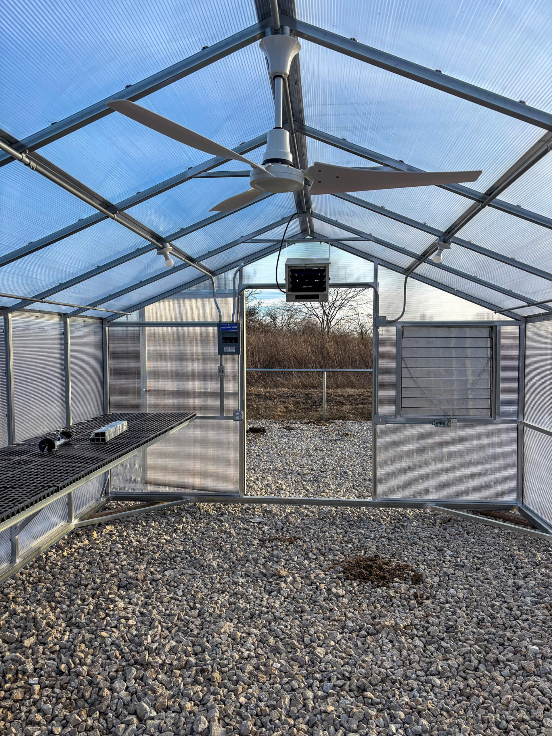 Sunny Crest Acres Versailles MO Interior of an empty greenhouse with transparent walls and ceiling, gravel floor, metal benches, ceiling fans, and an open door leading outside to a field.