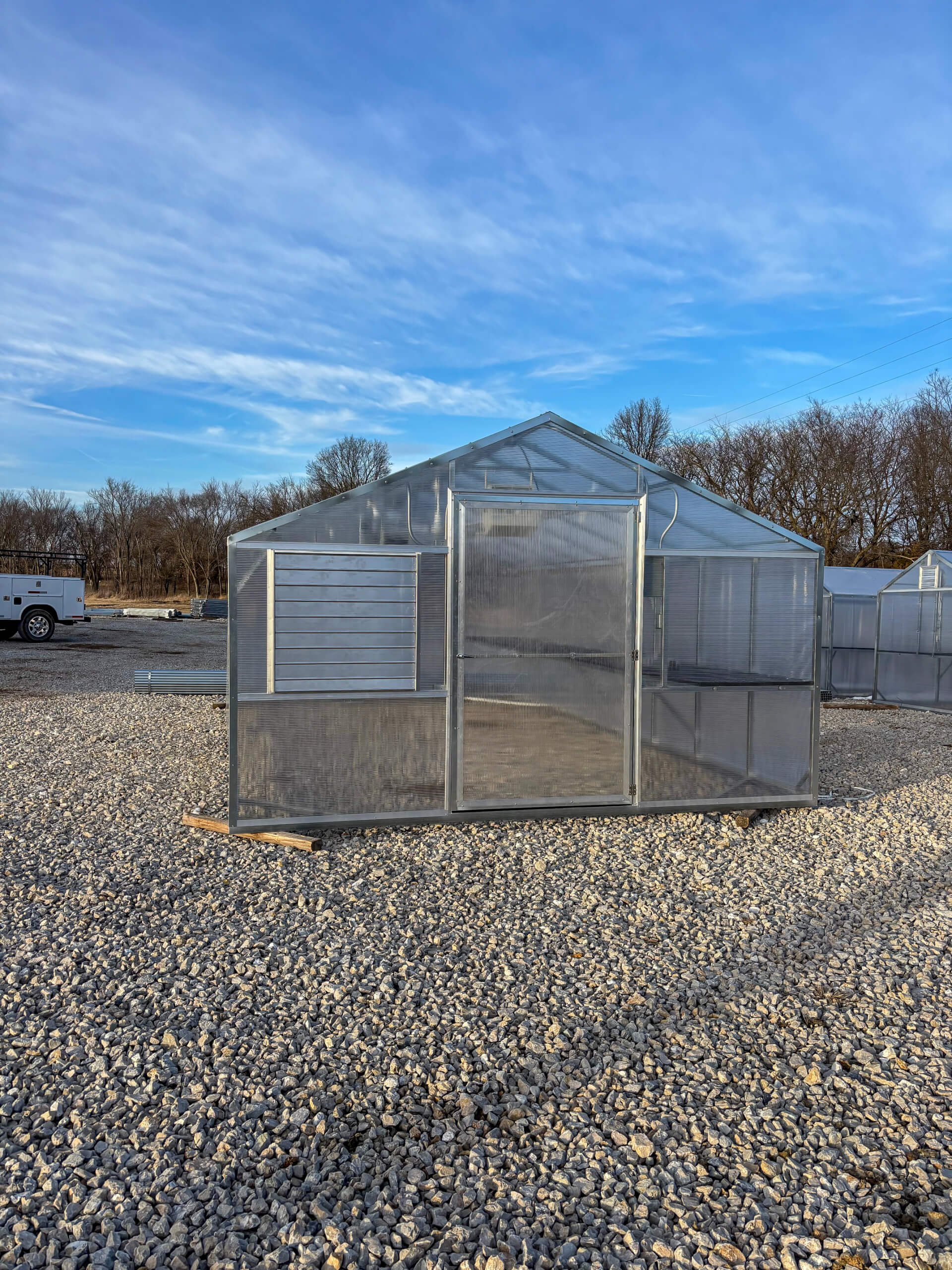 Sunny Crest Acres Versailles MO A small transparent greenhouse with a metal frame stands on a gravel lot under a blue sky, surrounded by bare trees and other structures in the background.