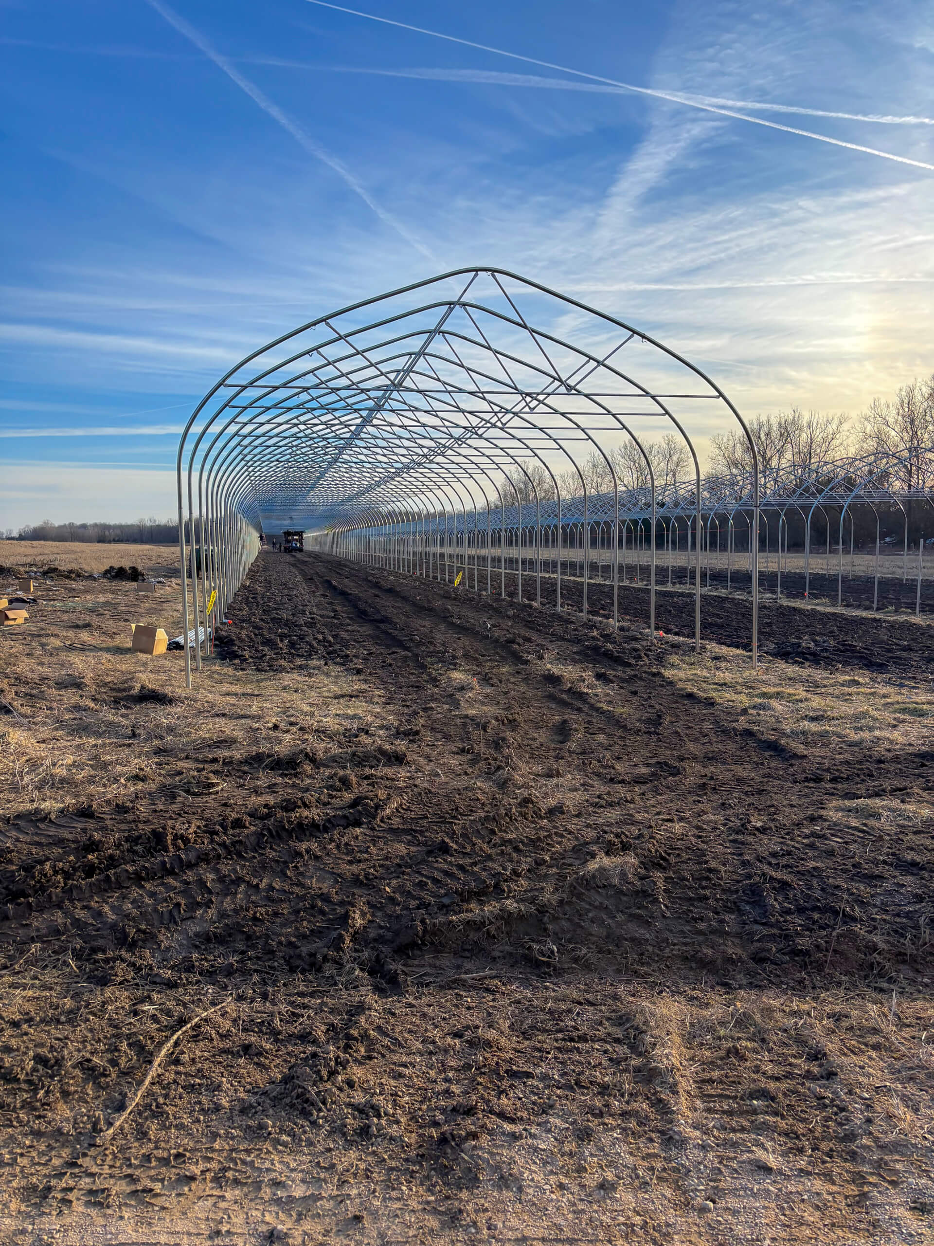 Sunny Crest Acres Versailles MO A partially constructed greenhouse frame stands on a dirt field under a blue sky with visible tire tracks leading inside.