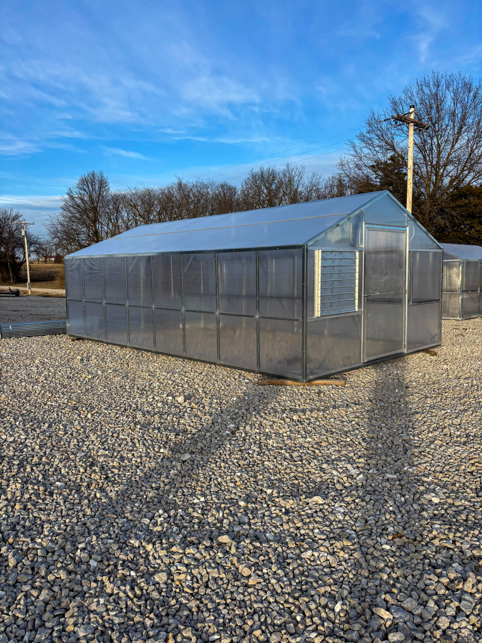 Sunny Crest Acres Versailles MO A transparent greenhouse stands on a gravel lot, surrounded by leafless trees under a clear blue sky.